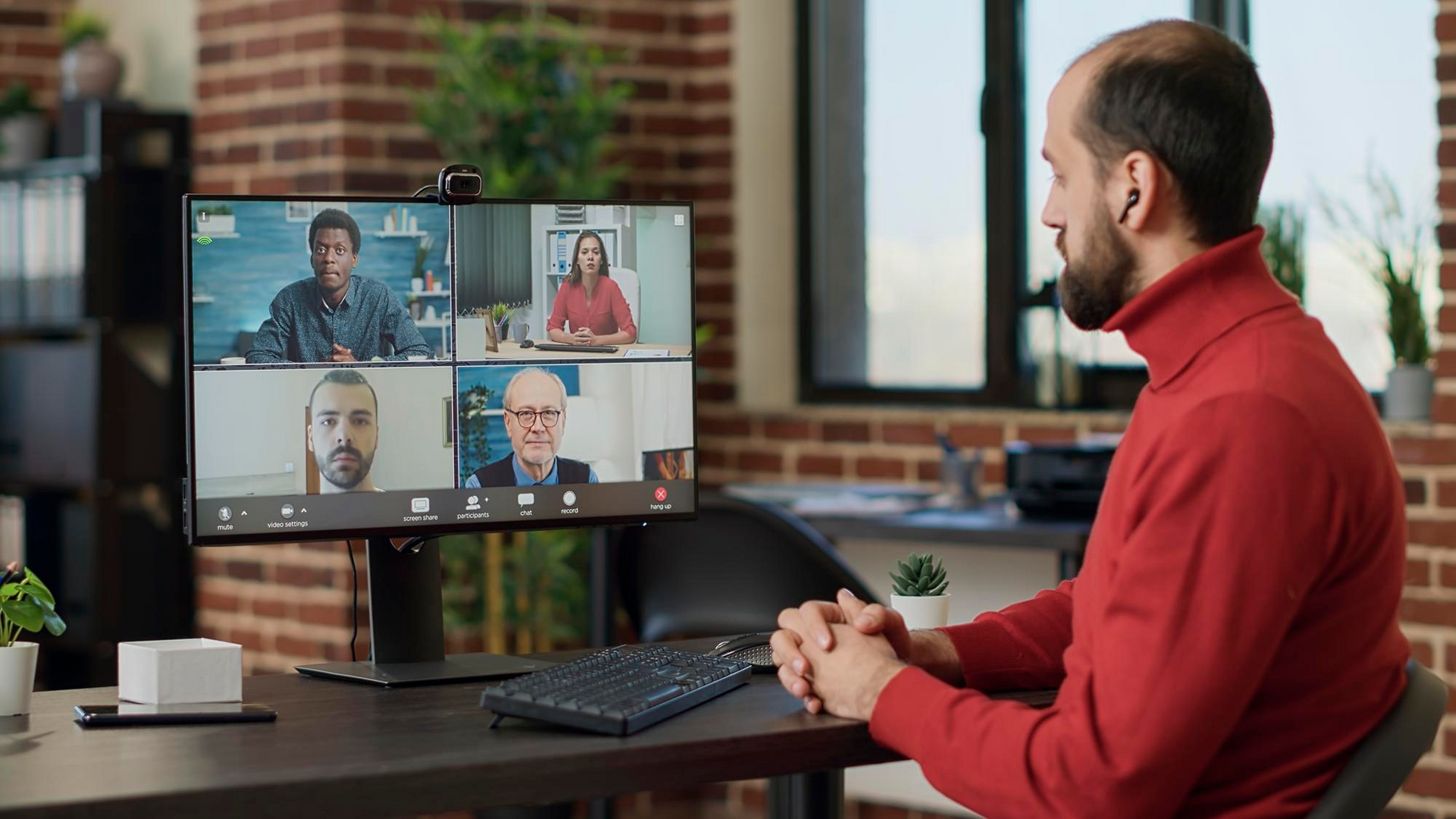 Person at desk in a video conference on a monitor, wearing a red sweater in a bright office.