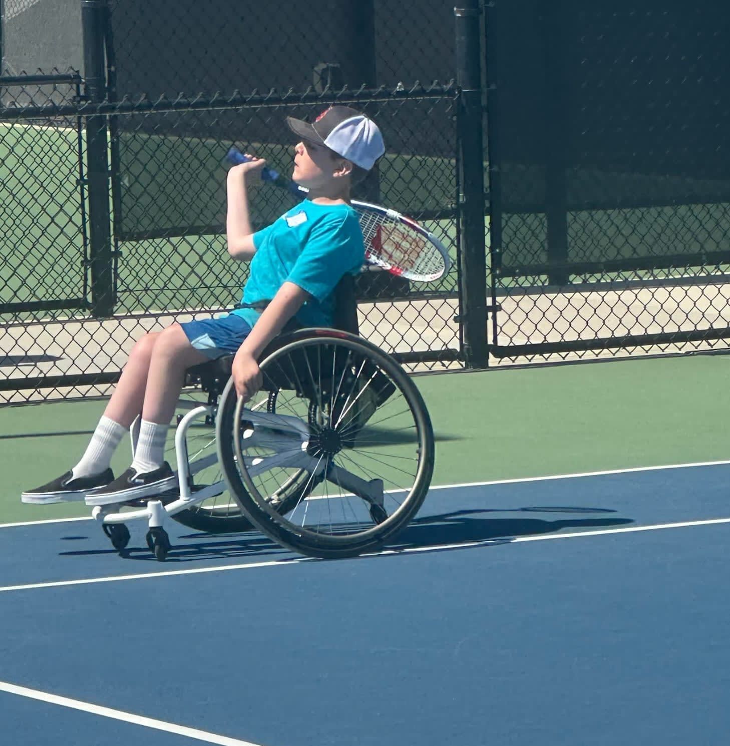 A young boy in a wheelchair drinking water on a tennis court