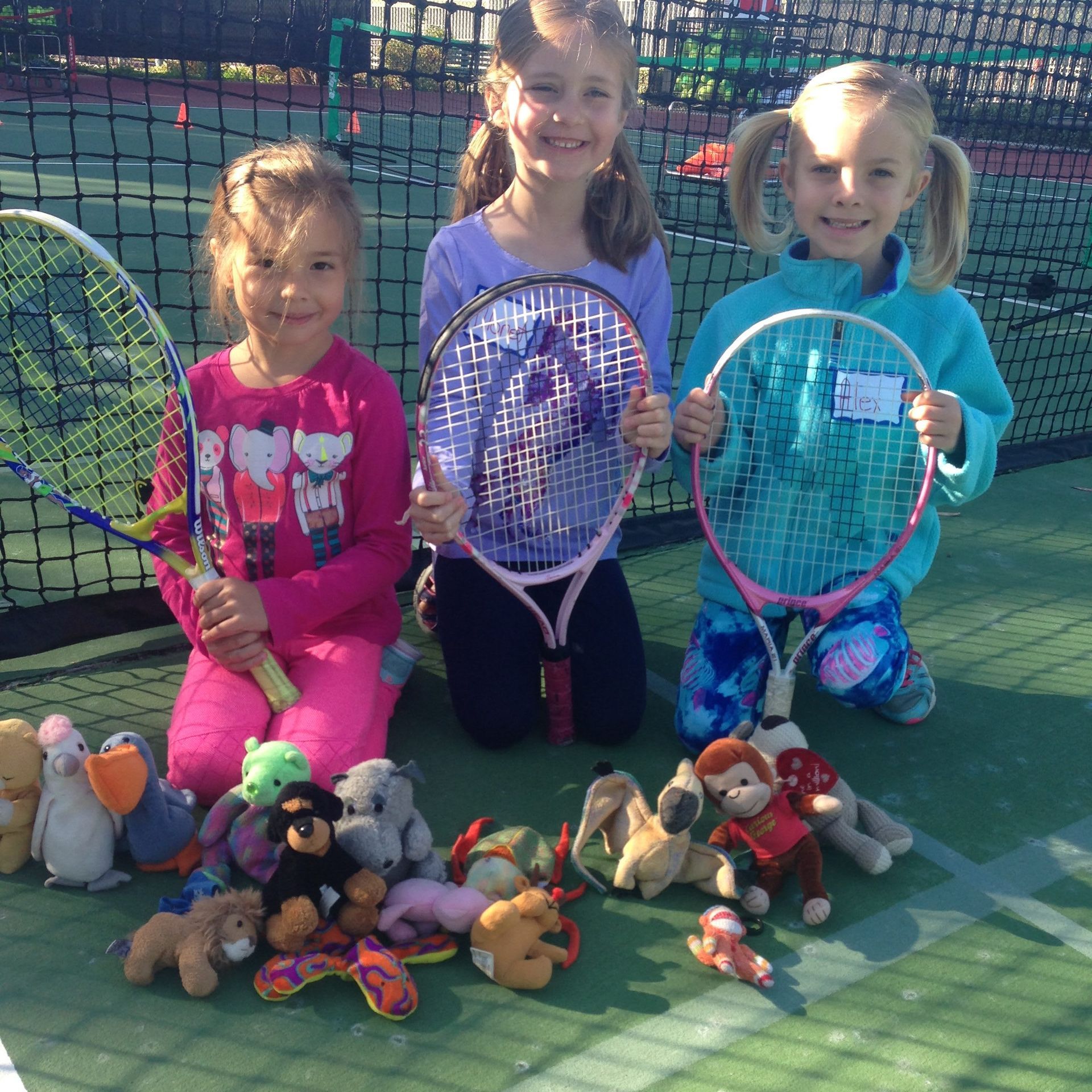 Three young girls are kneeling on a tennis court holding tennis rackets and stuffed animals