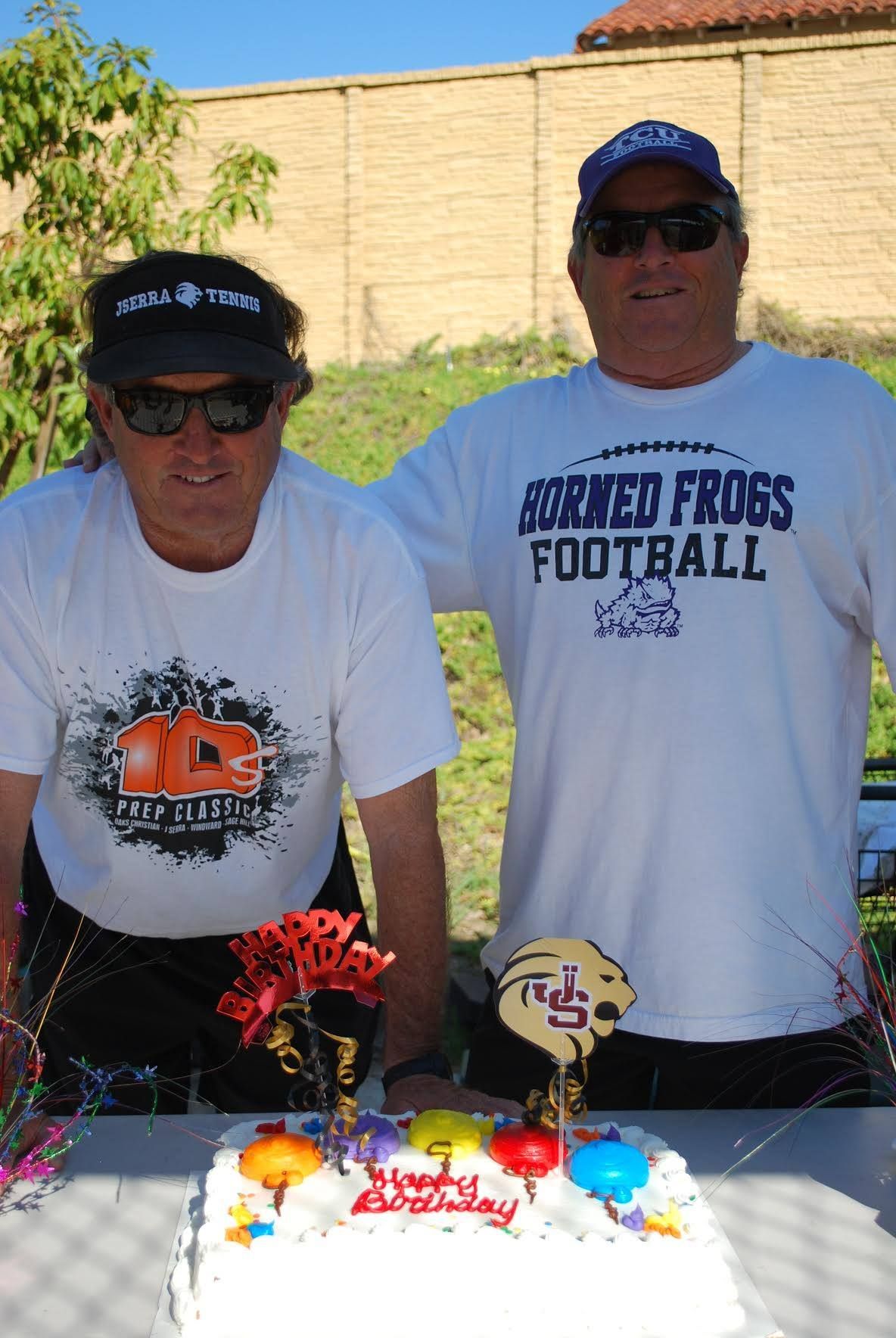 Two men wearing shirts that say turned frogs football are standing next to a cake