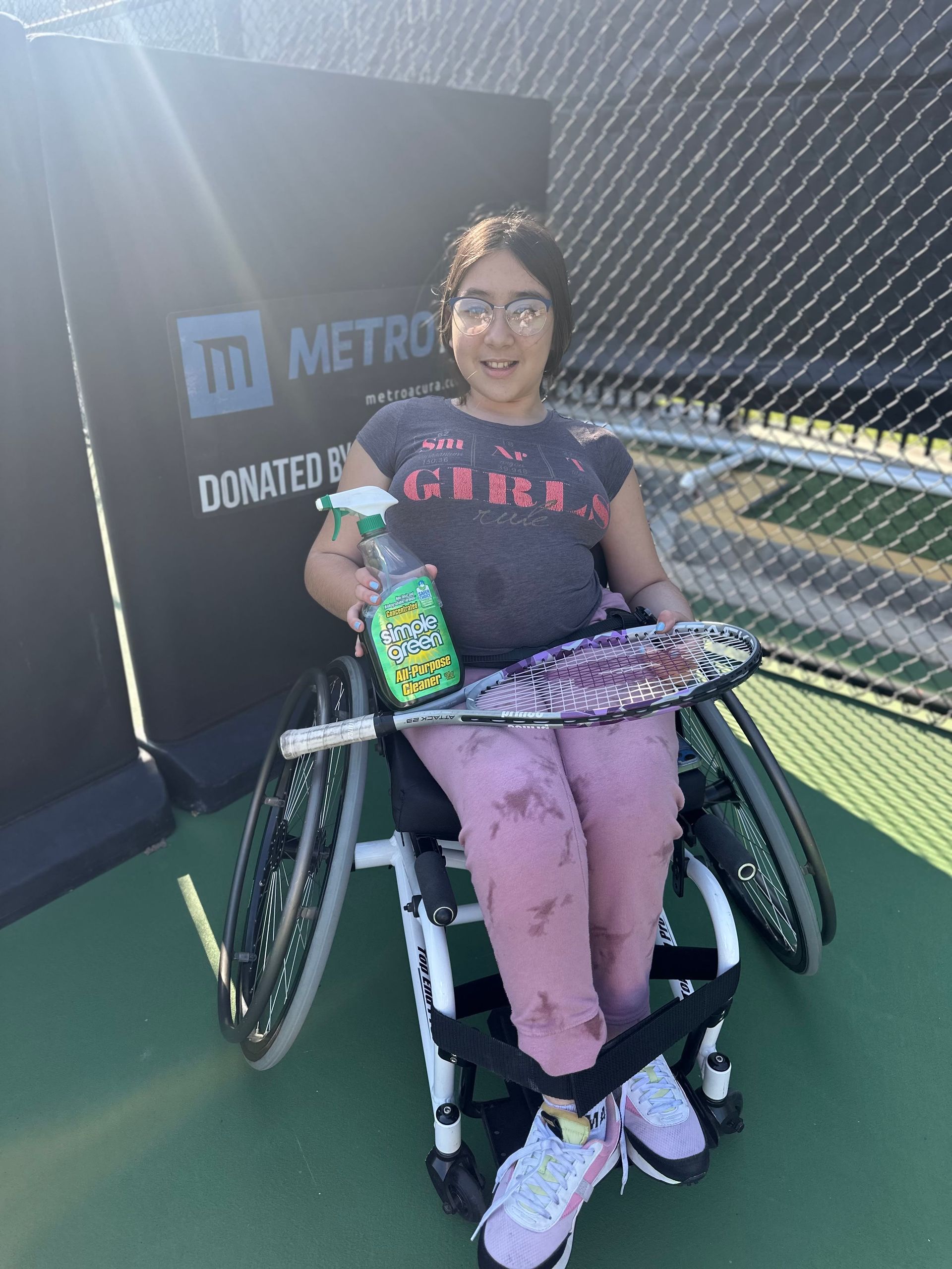A young girl in a wheelchair is holding a tennis racquet and a spray bottle.
