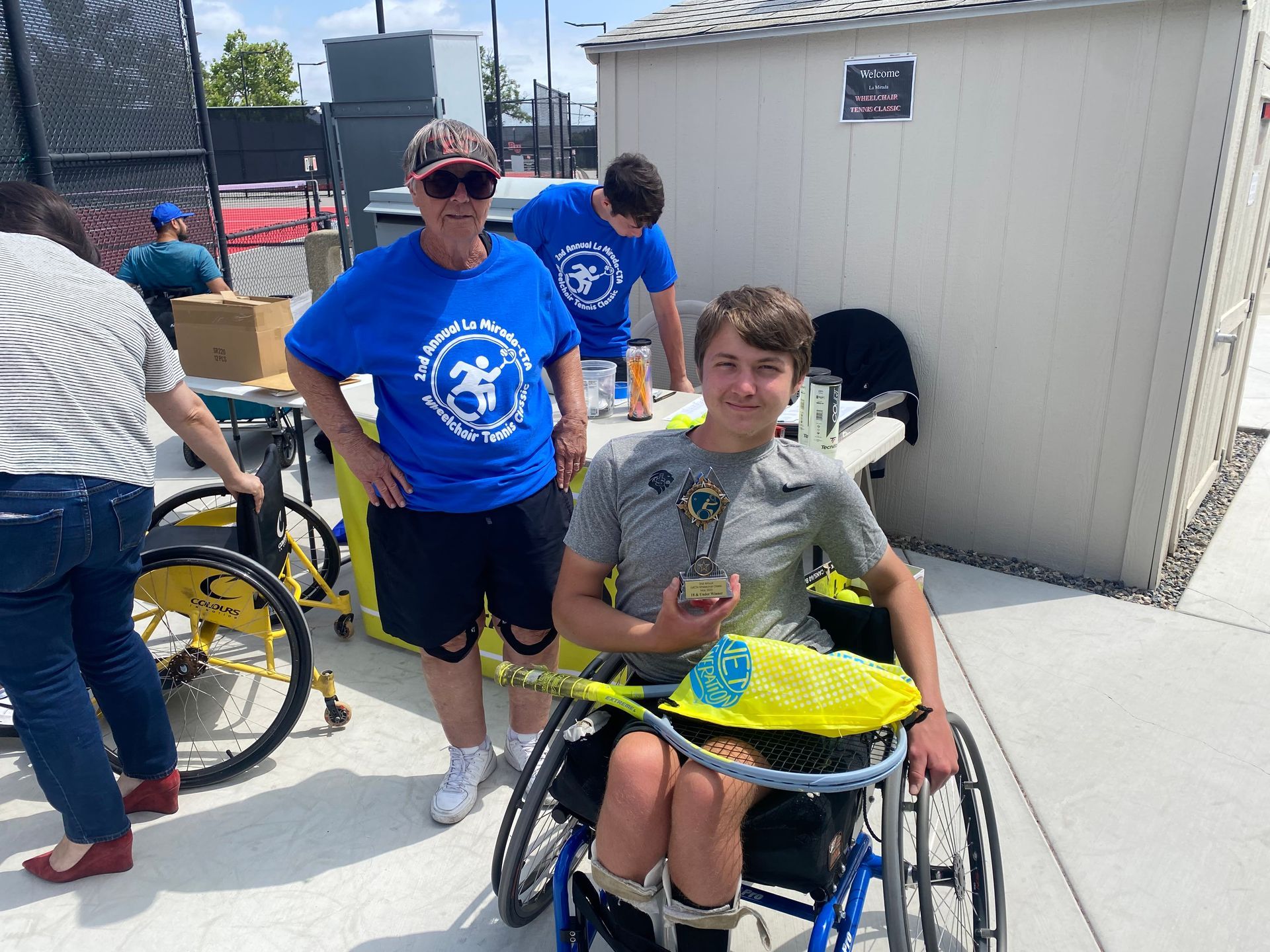 A man in a blue shirt is standing next to a boy in a wheelchair holding a tennis racket.