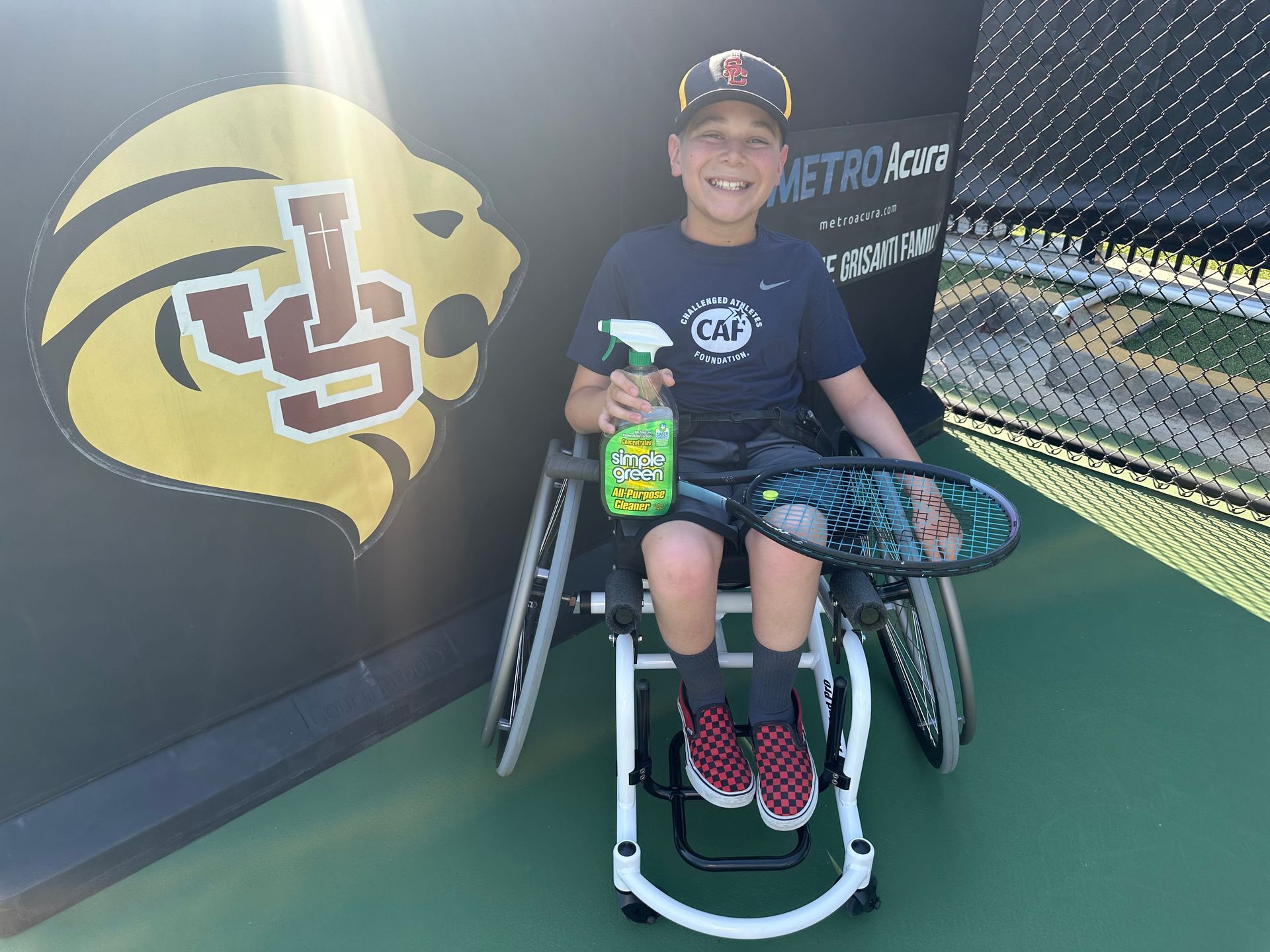 A young boy in a wheelchair is holding a spray bottle and a tennis racquet.