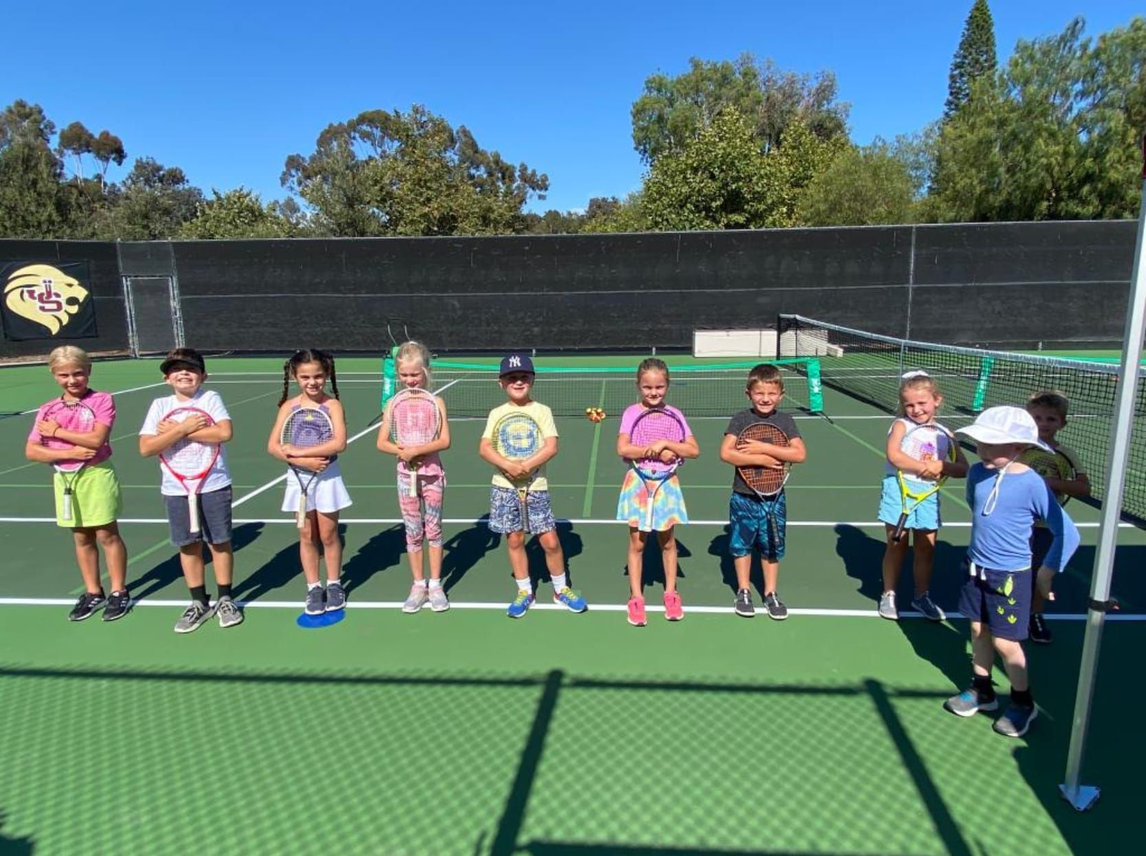 Three young girls are kneeling on a tennis court holding tennis rackets and stuffed animals