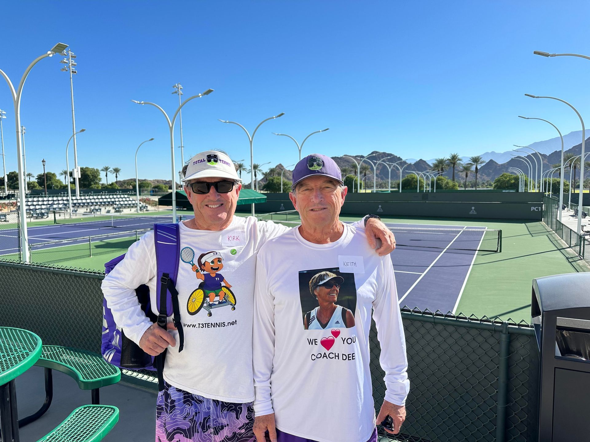 Two people standing on a tennis court wearing white long-sleeved shirts, one with a backpack, on a sunny day.