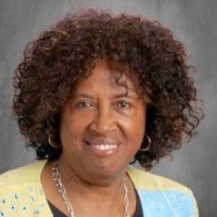 A woman with curly hair is wearing a yellow sweater and a necklace.