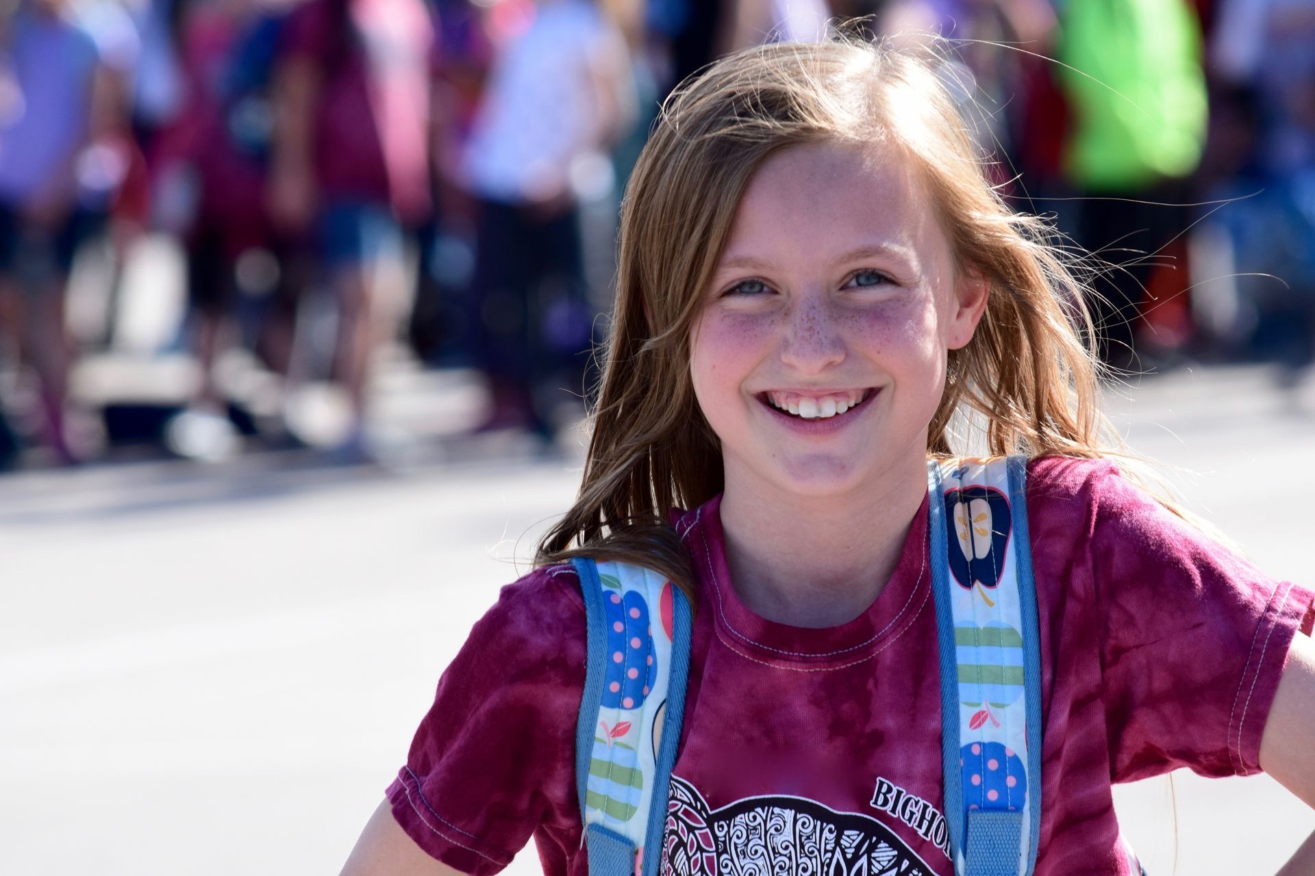 A young girl wearing a tie dye shirt and a backpack is smiling.