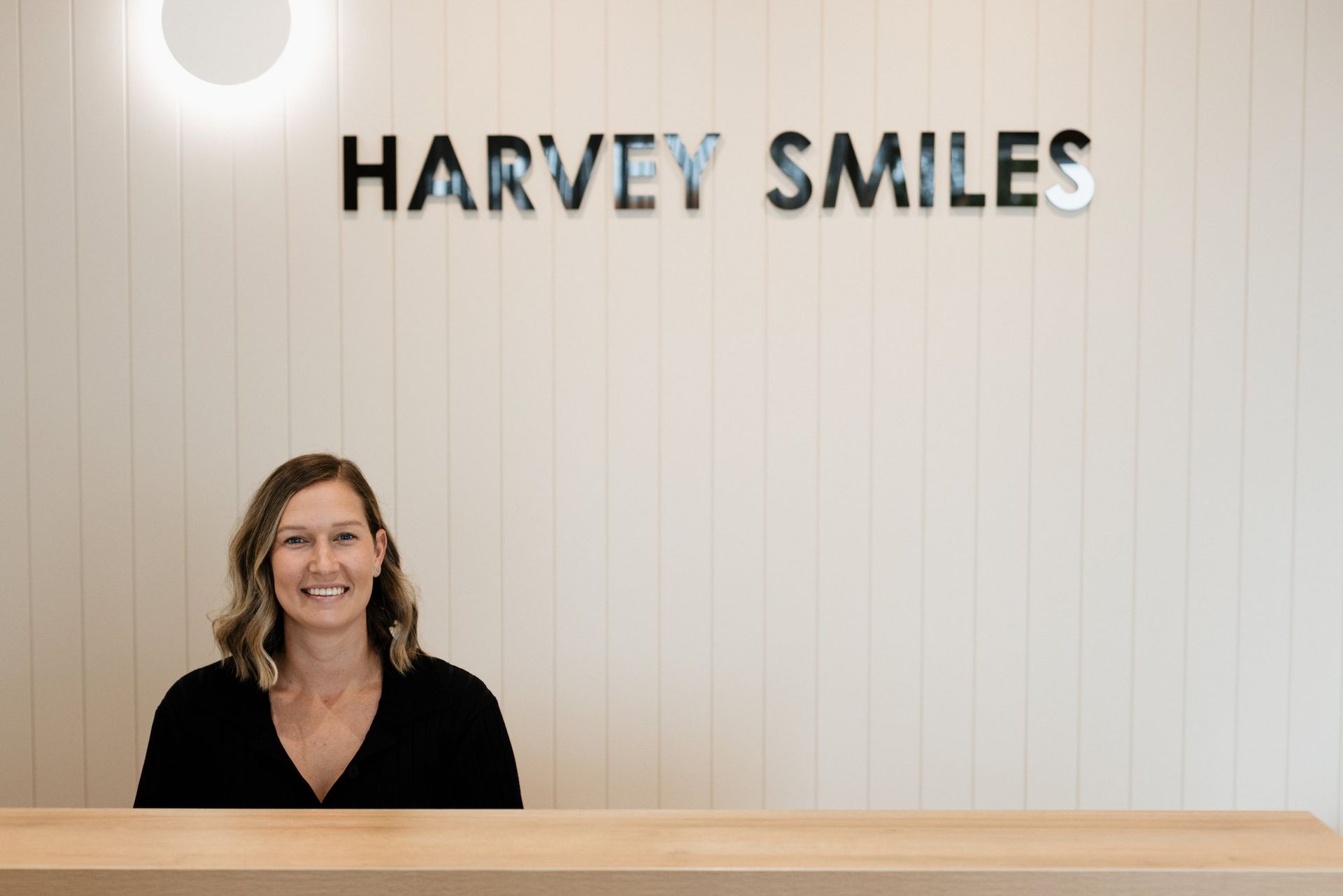 A Woman Is Sitting At A Desk In Front Of A Sign That Says Harvey Smiles — Harvey Smiles Denture Clinic In Burleigh Heads, QLD