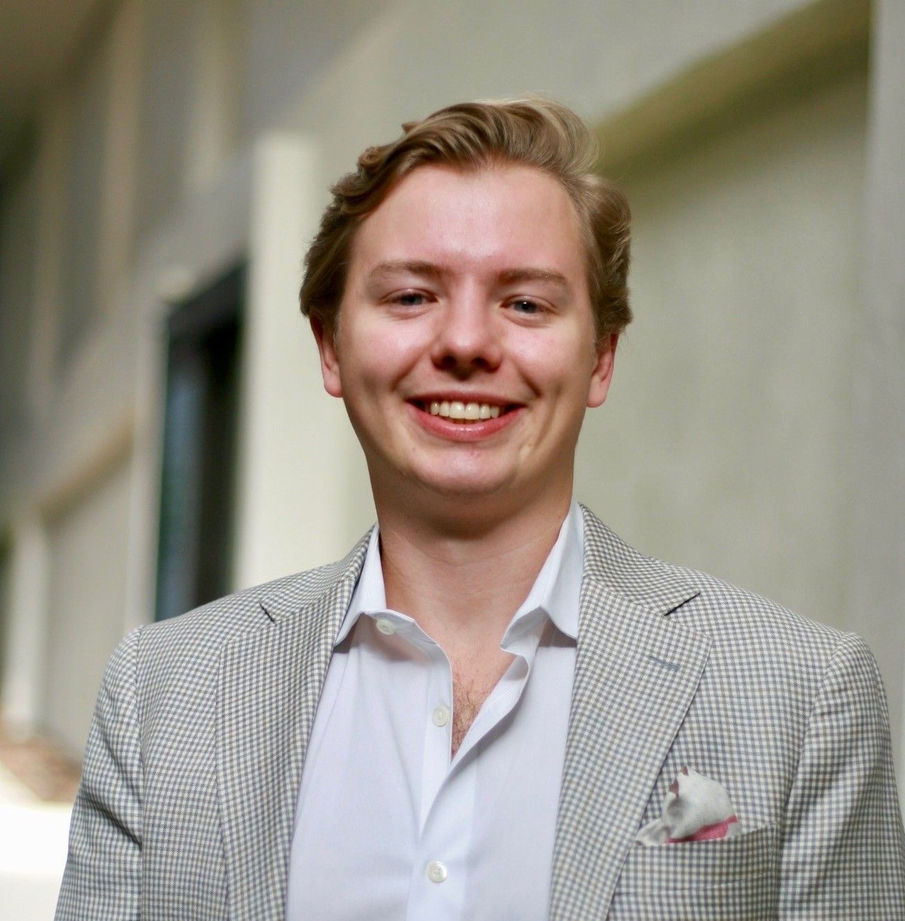 Man in a light-colored blazer smiling at the camera, leaning against a building.
