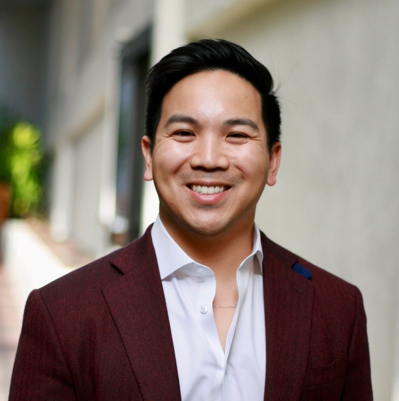 Man in burgundy blazer, white shirt, smiling outdoors.