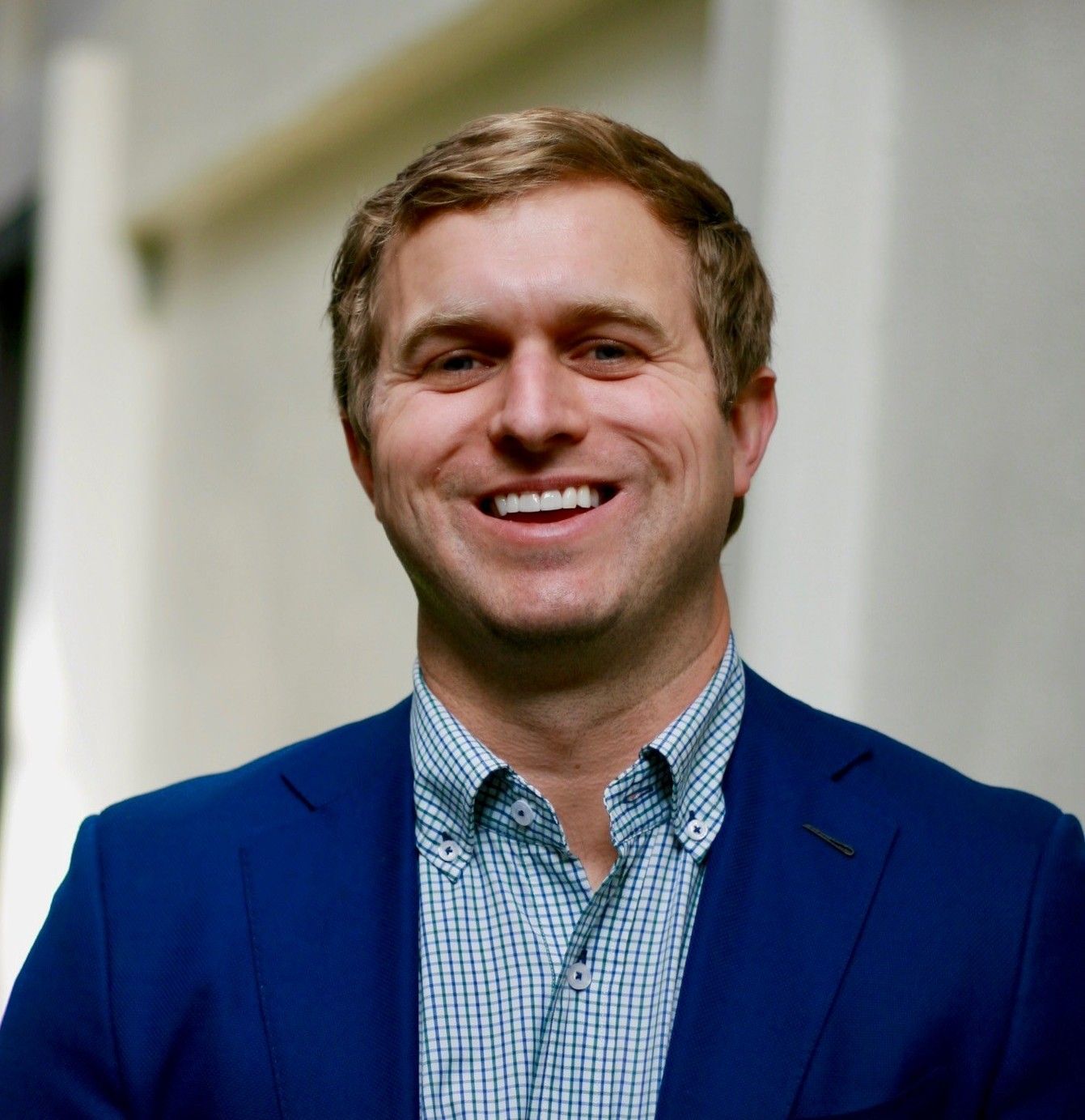 Man in blue blazer smiling, light-colored hair, button-down shirt, neutral background.