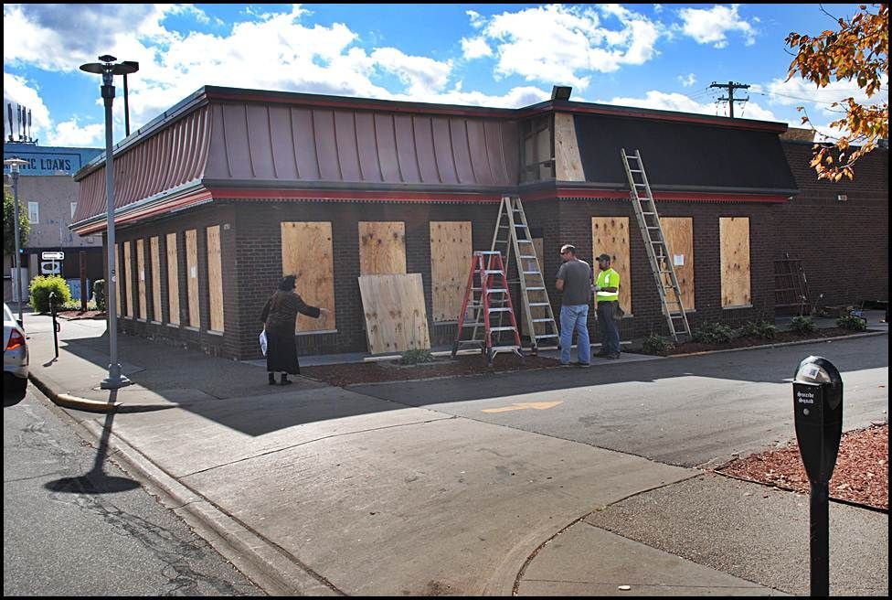 Building with boarded-up windows, construction workers, ladders, and a person walking on a sidewalk.