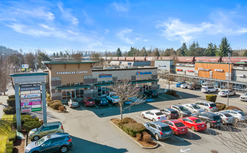 Exterior view of a shopping center with various stores, parked cars, and a blue sky.