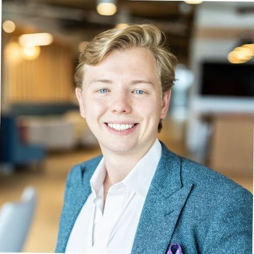 Smiling man with blonde hair, in a blazer and white shirt, in a modern office.
