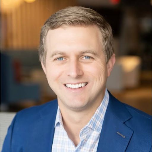 Smiling man in blue blazer and light blue plaid shirt, blurred office background.