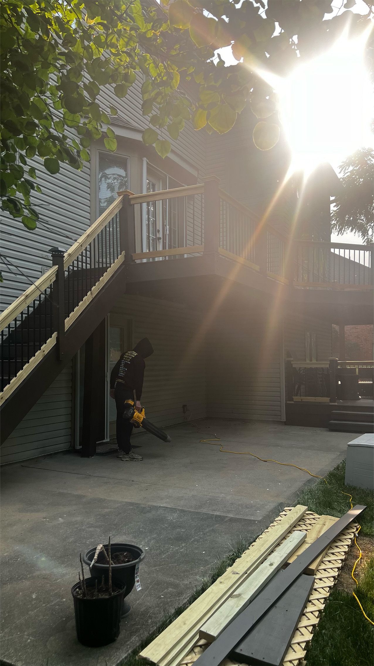 A man is working on a deck in front of a house with the sun shining through the trees.