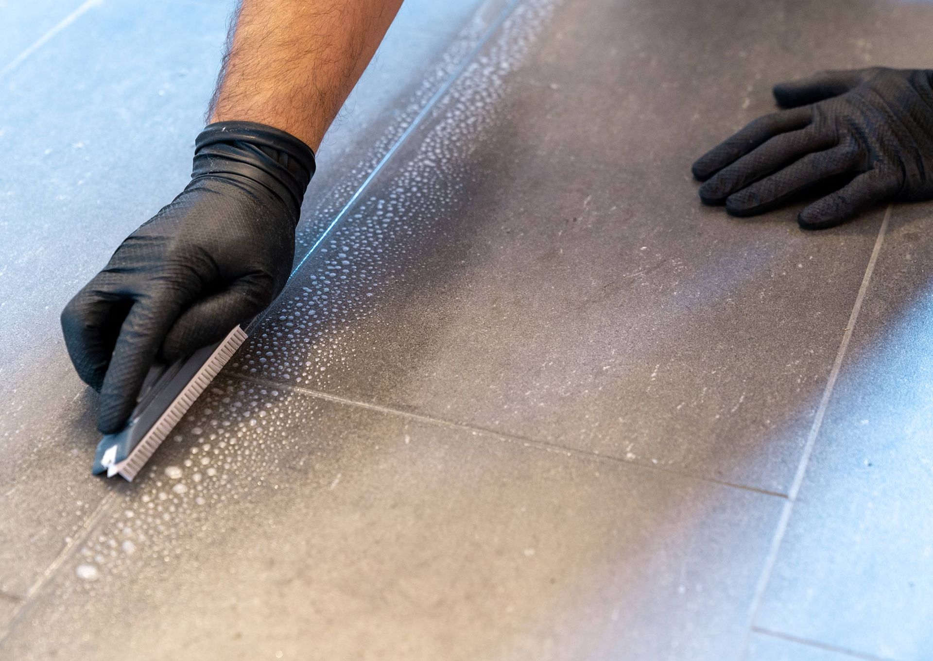 A person wearing black gloves is cleaning a tile floor with a brush.