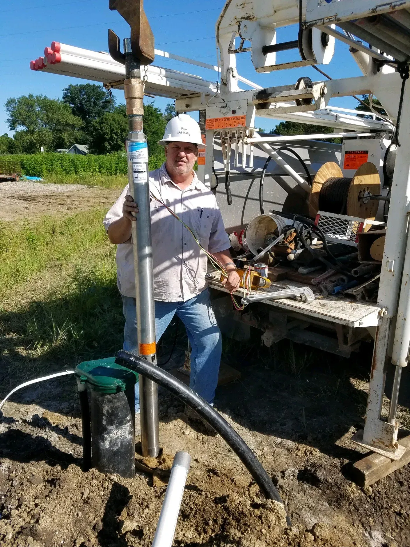 Man in hard hat holds a metal pipe near a well drilling rig. Outdoors, sunny day.