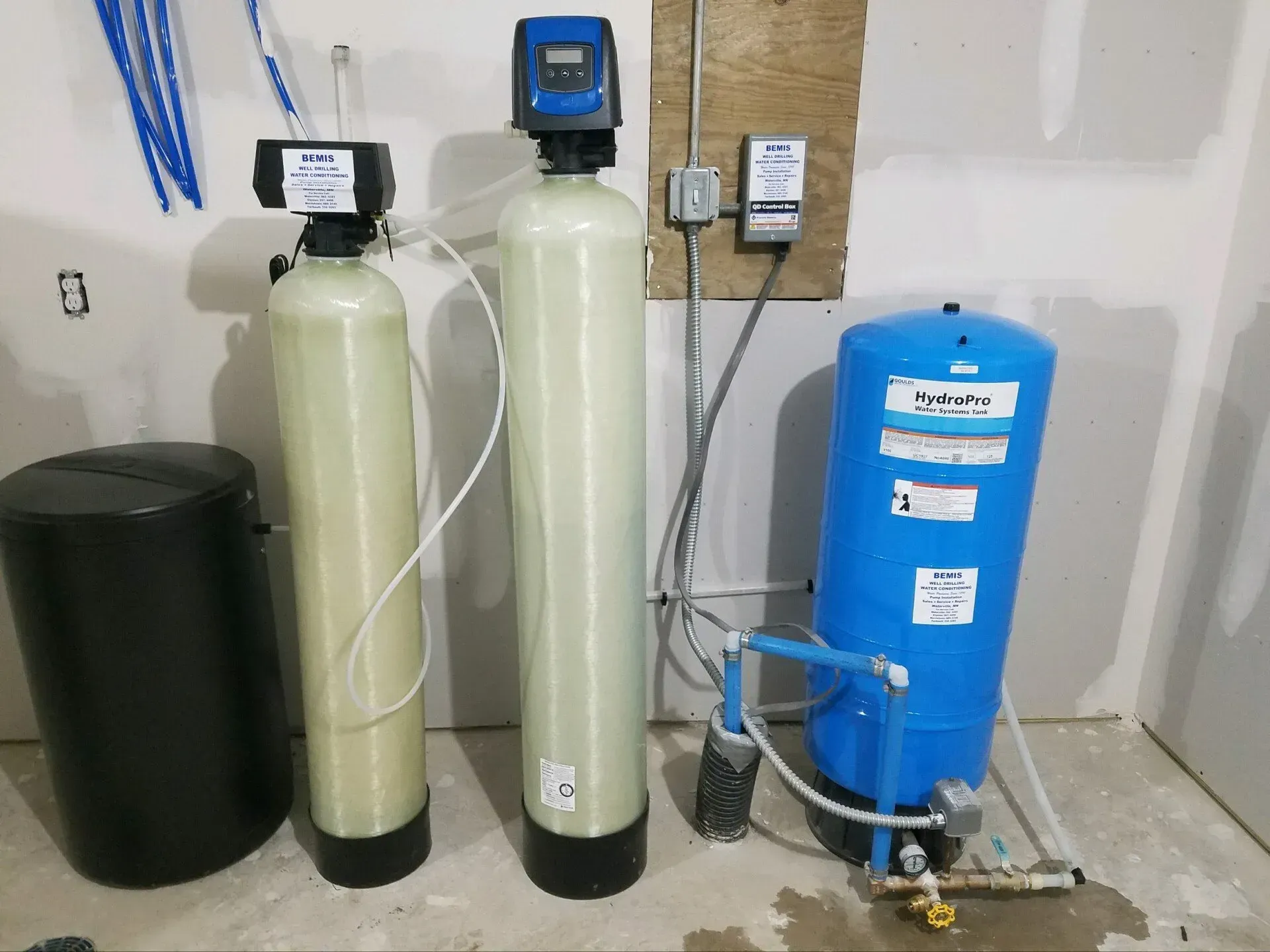 Water softener and pressure tank system in a utility room.  Tanks are beige and blue. Black tank on the left.