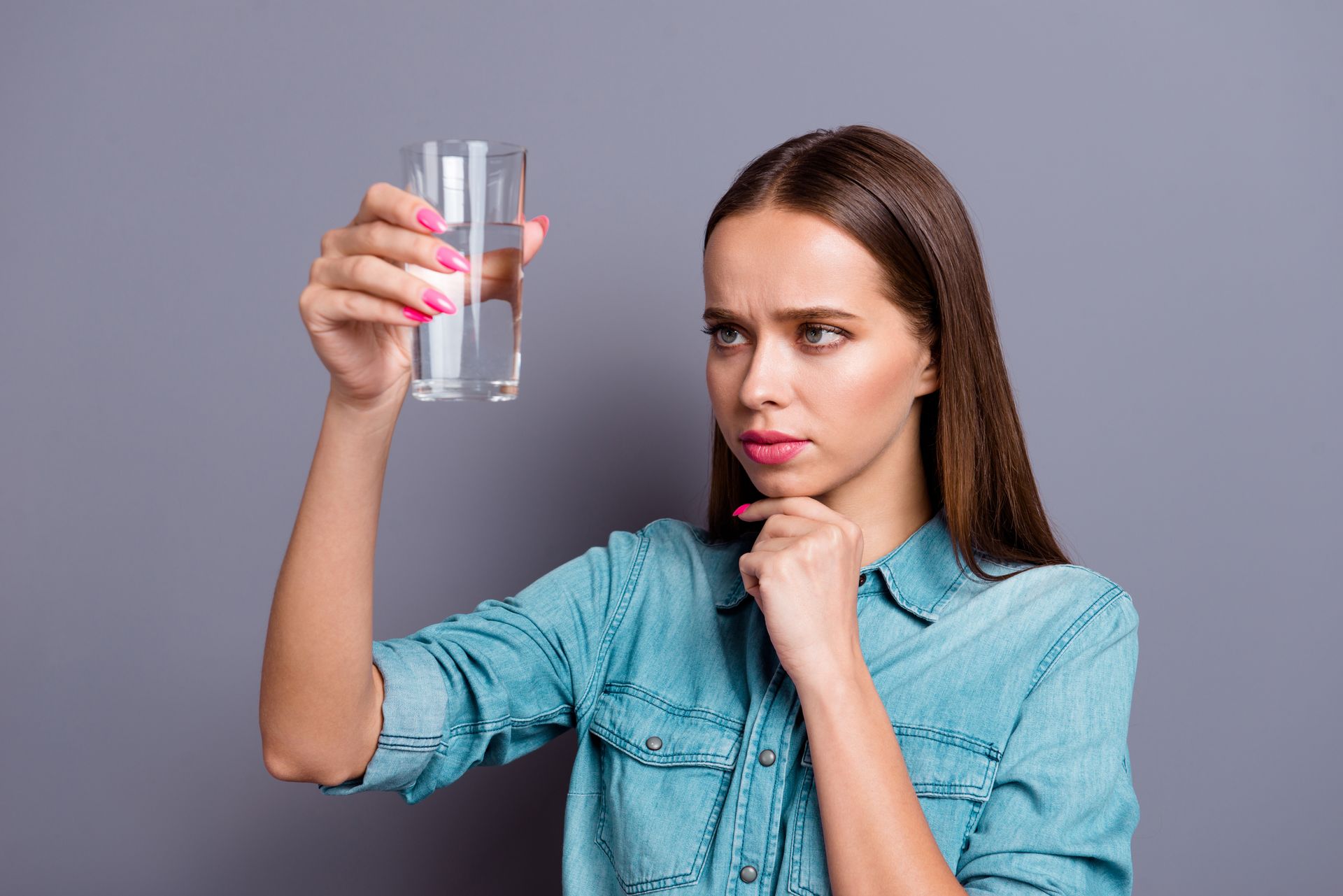 Woman with a serious expression examining a glass of water, against a gray backdrop.