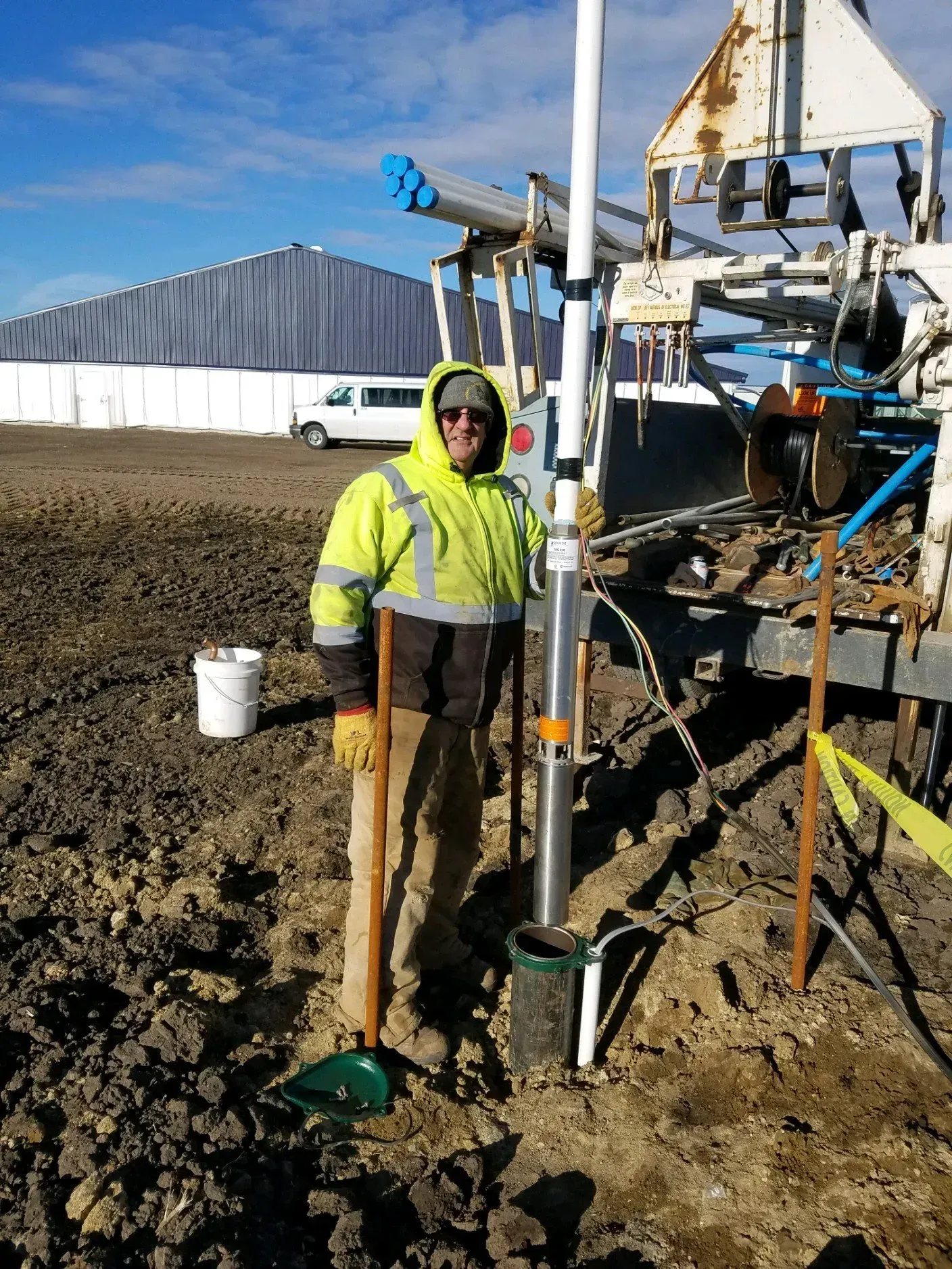 Man in high-vis vest by well casing on a muddy construction site. White building and truck in background.