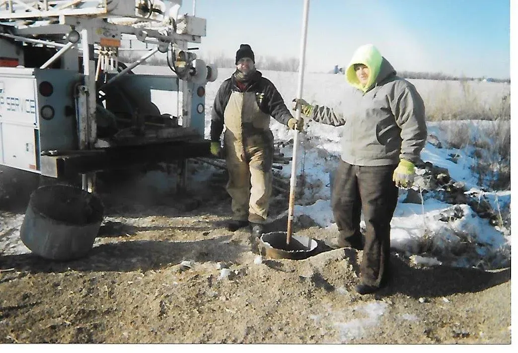 Two people at a drilling site, snowy ground. One holds a rod, the other stands near machinery, soil.