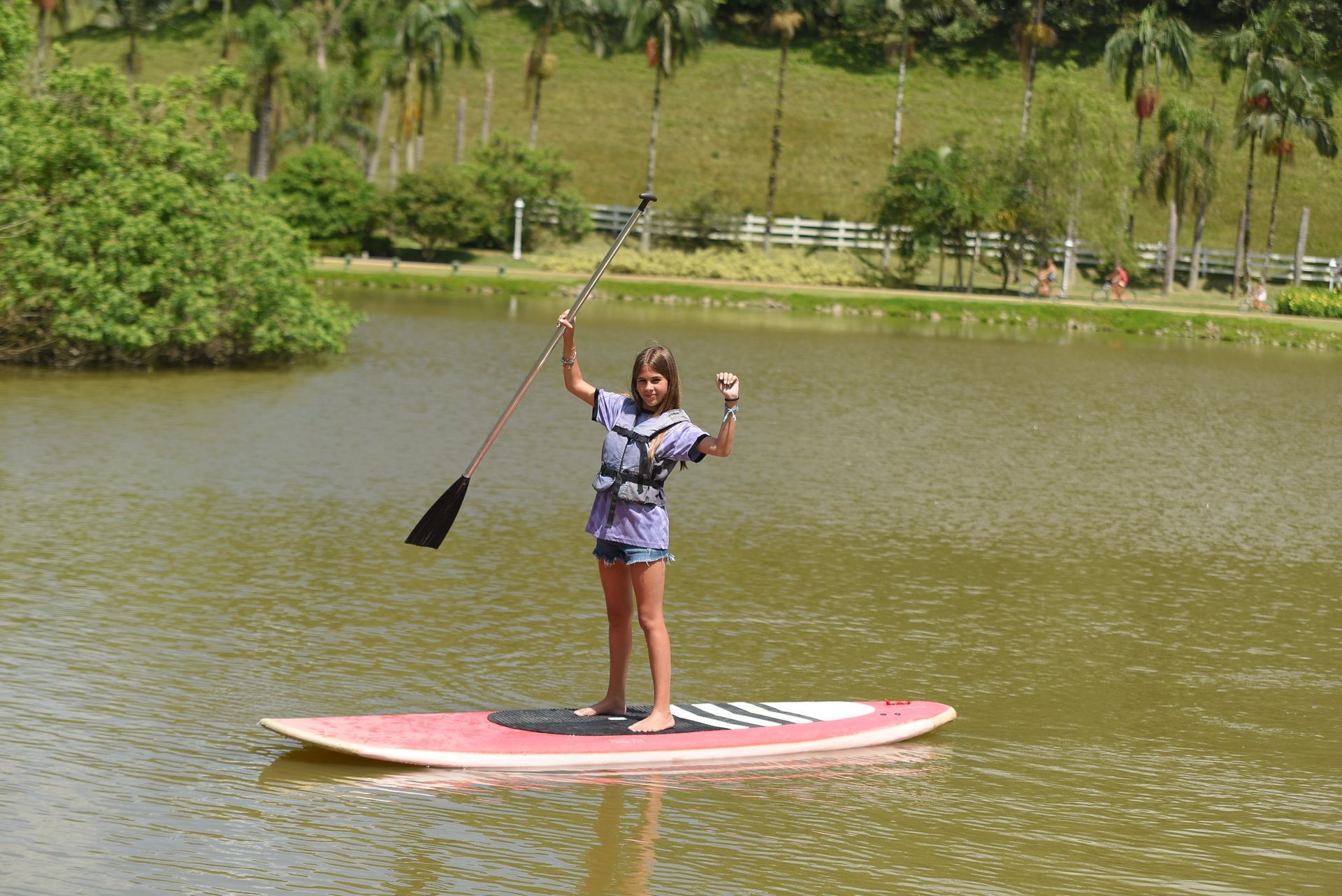 Pessoa praticando stand-up paddle em um lago, segurando um remo e com um braço levantado em um parque arborizado.