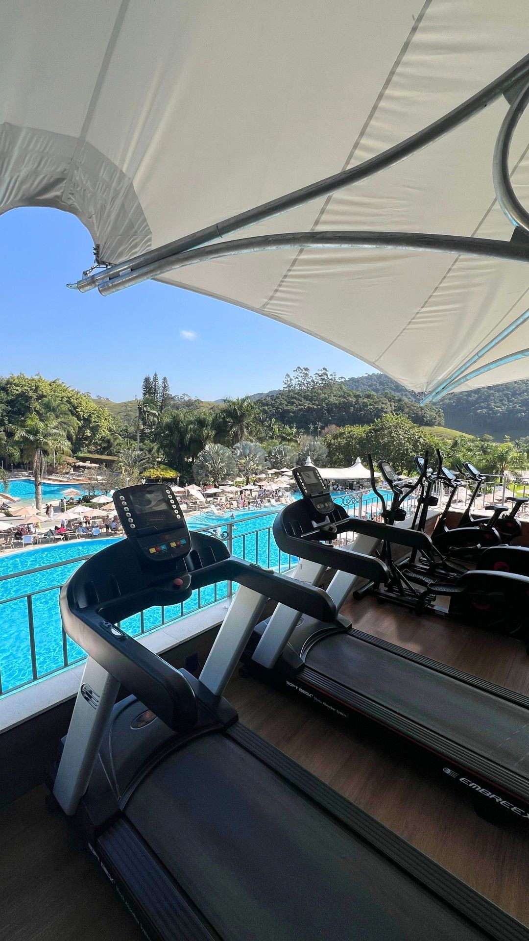 Área de descanso sombreada à beira da piscina com bicicletas ergométricas, água azul e árvores sob um céu claro.