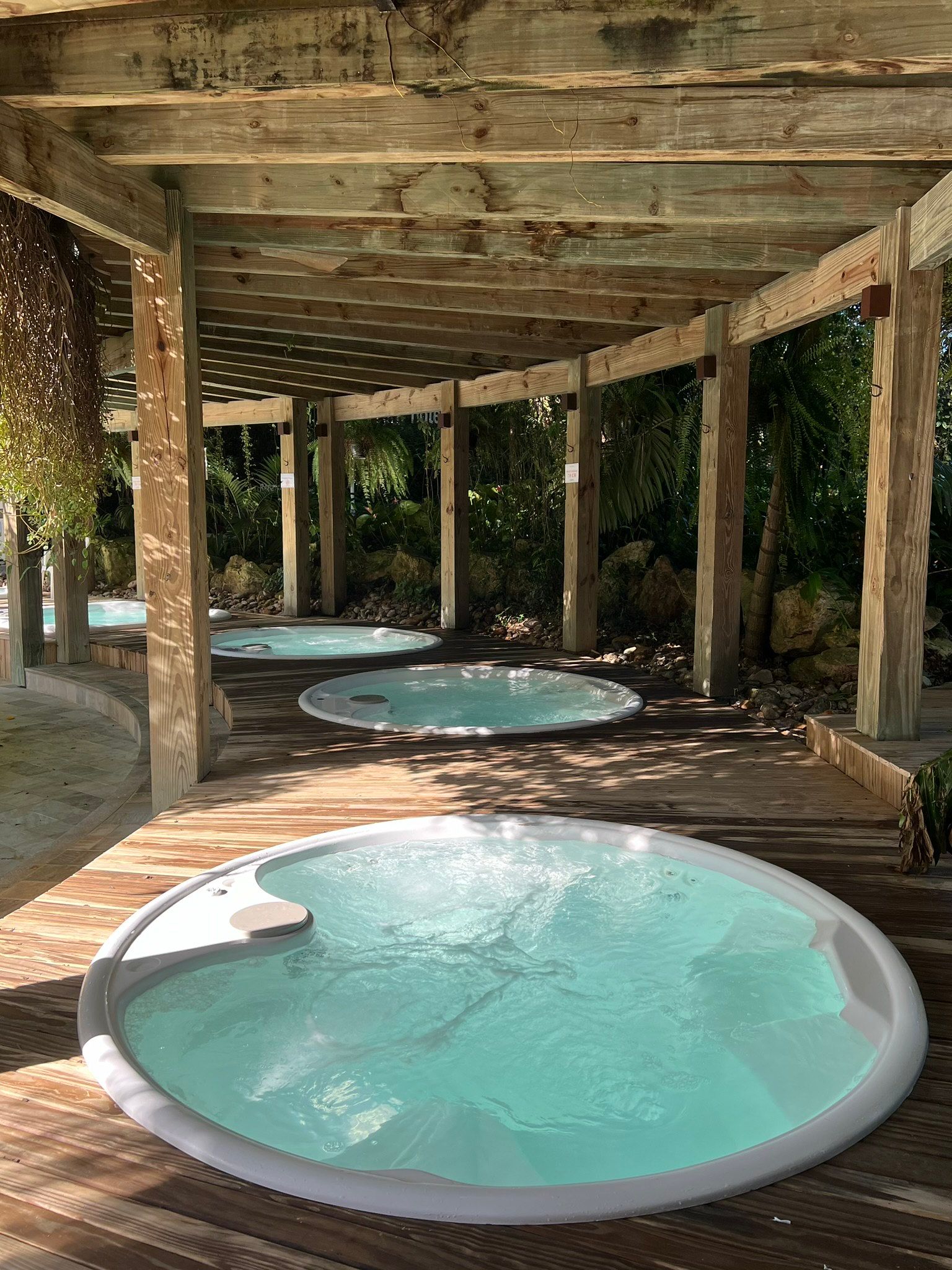 Três banheiras de hidromassagem redondas sob uma pérgola rústica de madeira em uma área de spa externa sombreada.