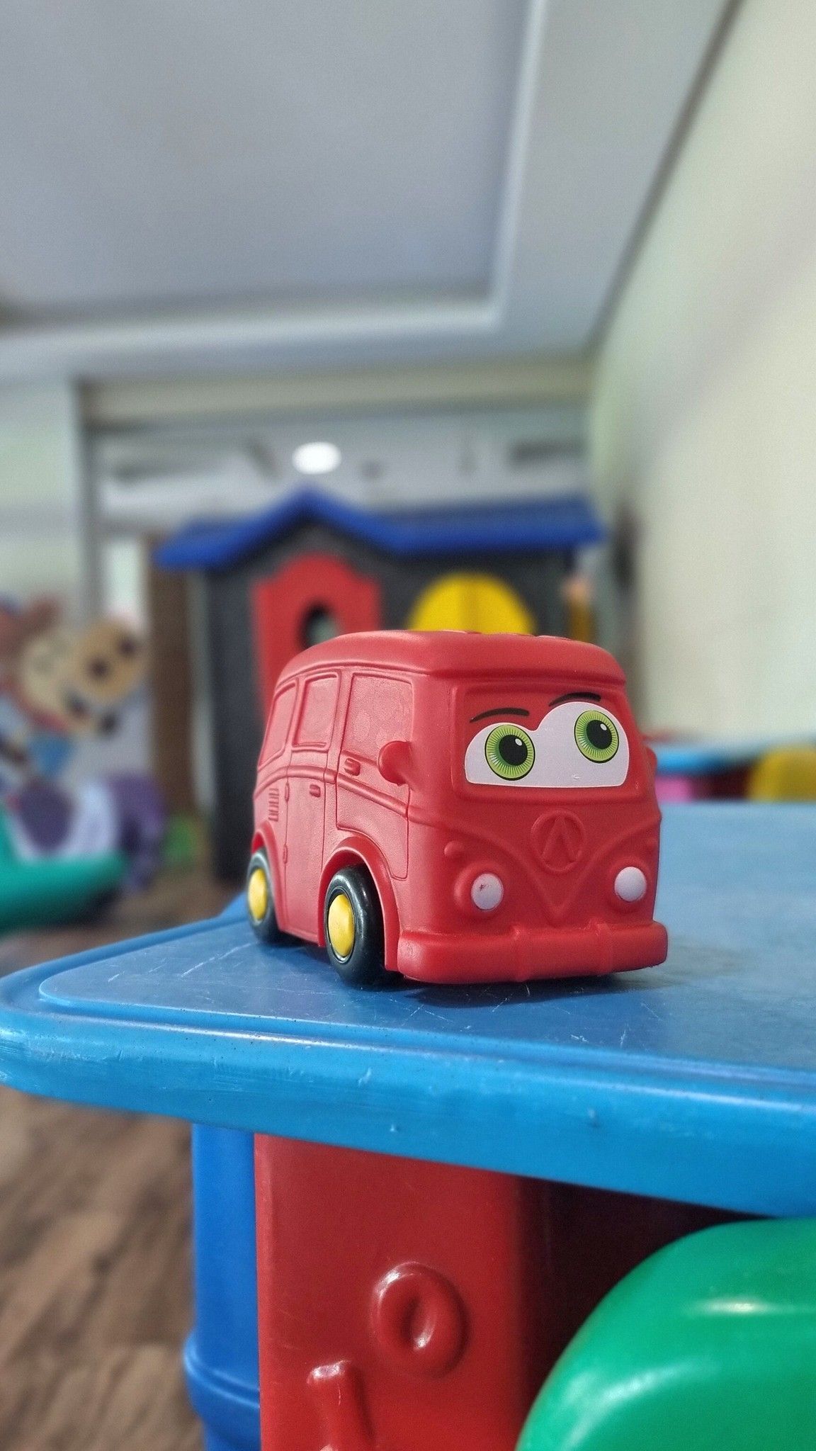 Ônibus de brinquedo vermelho sobre uma mesa de atividades azul no quarto de uma criança, com brinquedos desfocados ao fundo.