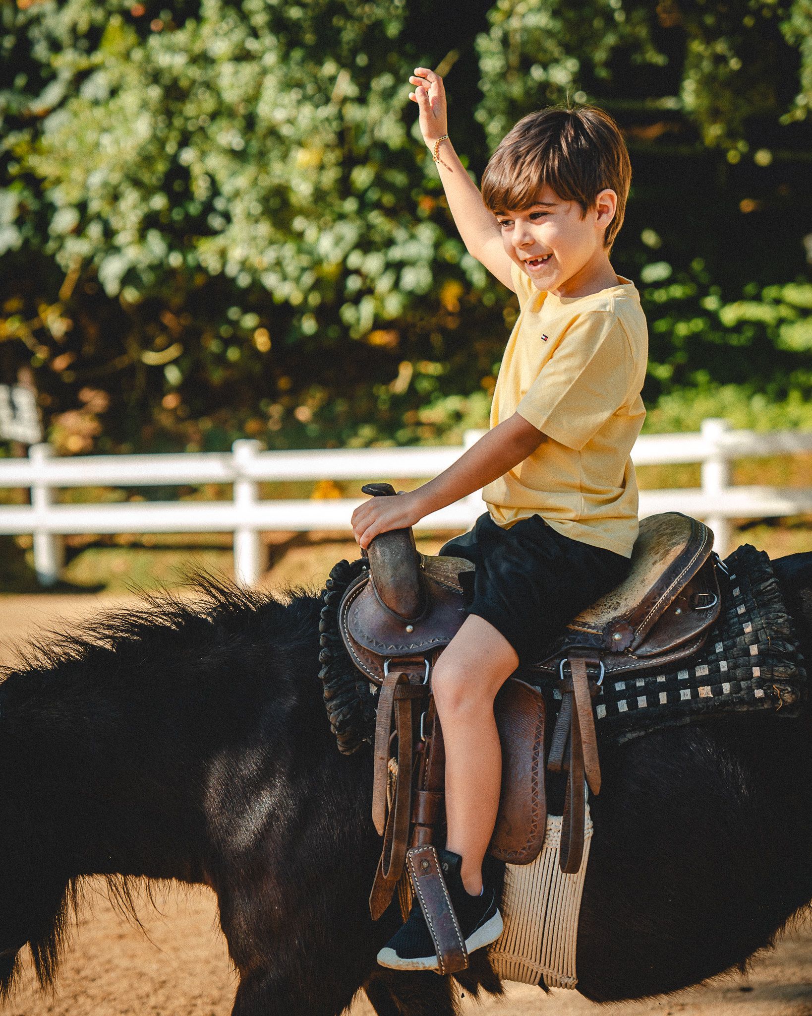 Criança a cavalo, sorrindo e com um braço levantado em uma área ensolarada para equitação ao ar livre.