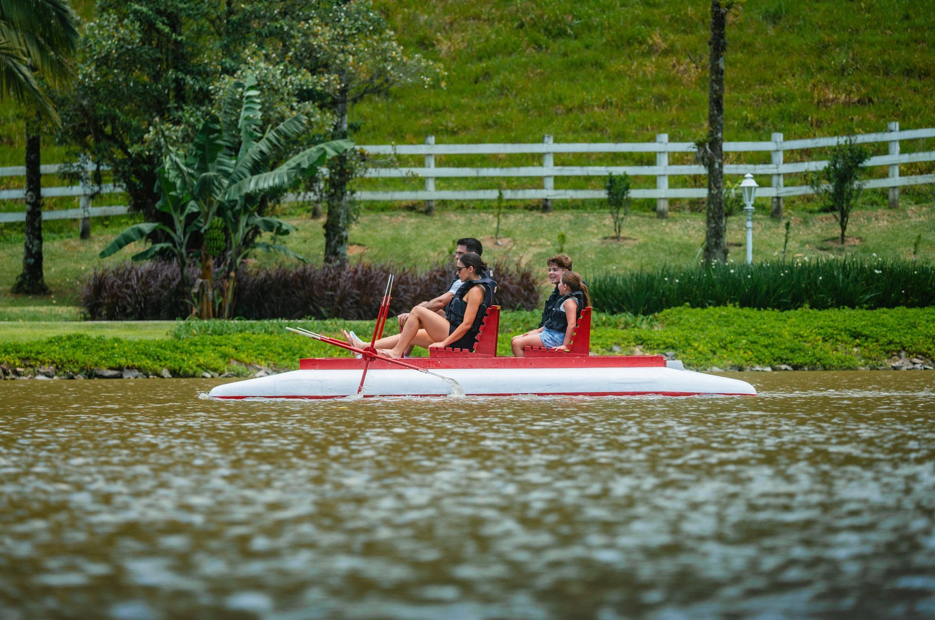 Duas pessoas remando em uma canoa vermelha em um lago com uma cerca branca e árvores verdes ao fundo.