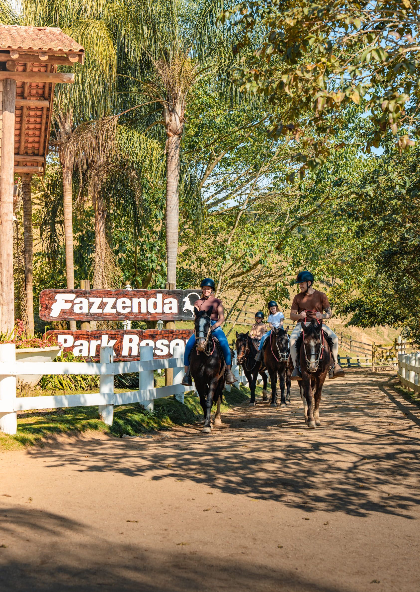 Duas pessoas a cavalo cavalgam em direção à câmera na Fazenda Karl Rosse, com uma placa e árvores próximas.