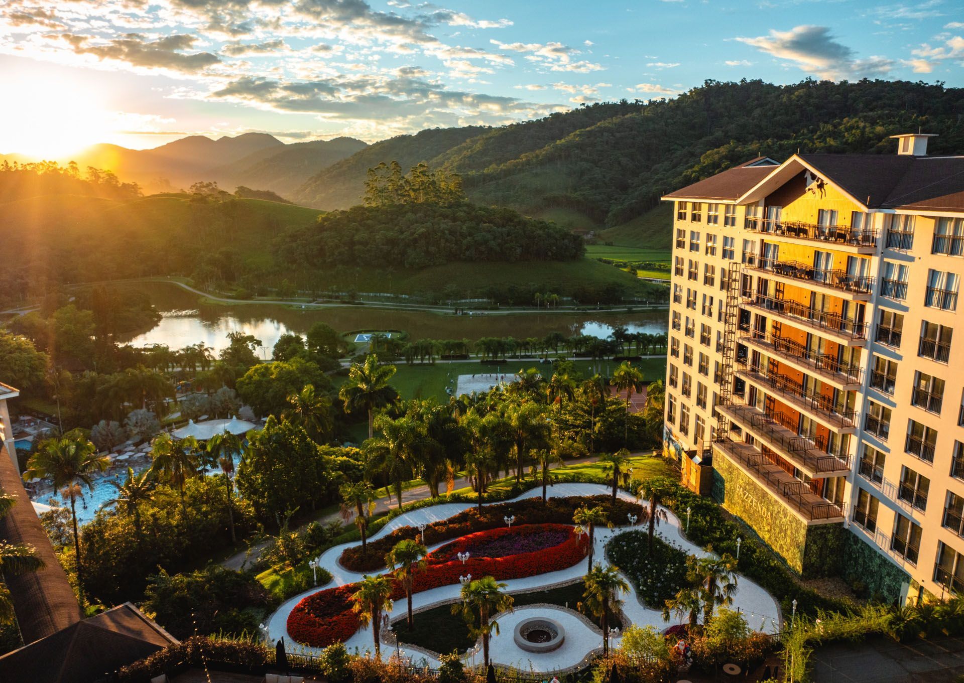 Pôr do sol sobre um resort de montanha com piscina, jardins e um edifício de hotel à beira do lago.