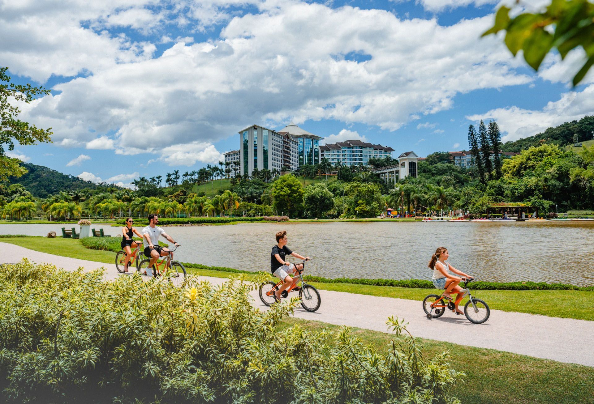 Pessoas andando de bicicleta em uma ciclovia à beira do rio, com céu nublado e prédios modernos ao fundo.