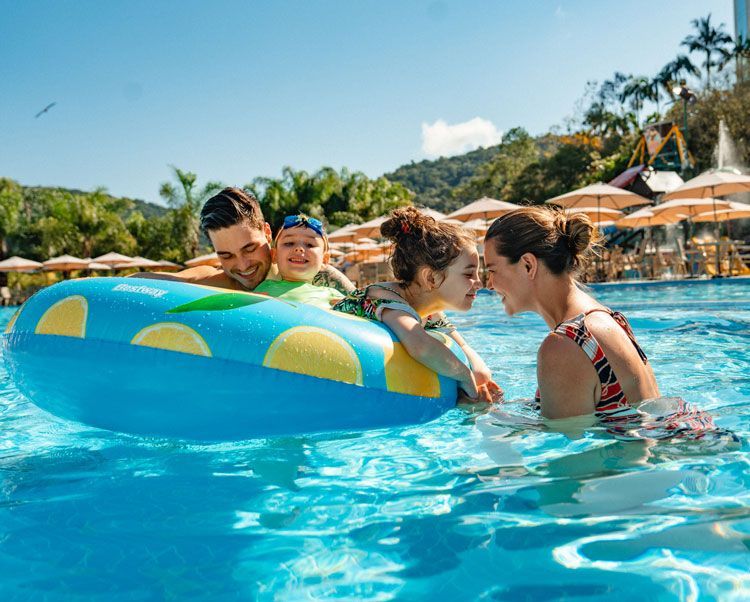 Família brincando em uma piscina com uma boia inflável colorida em um resort tropical ensolarado.