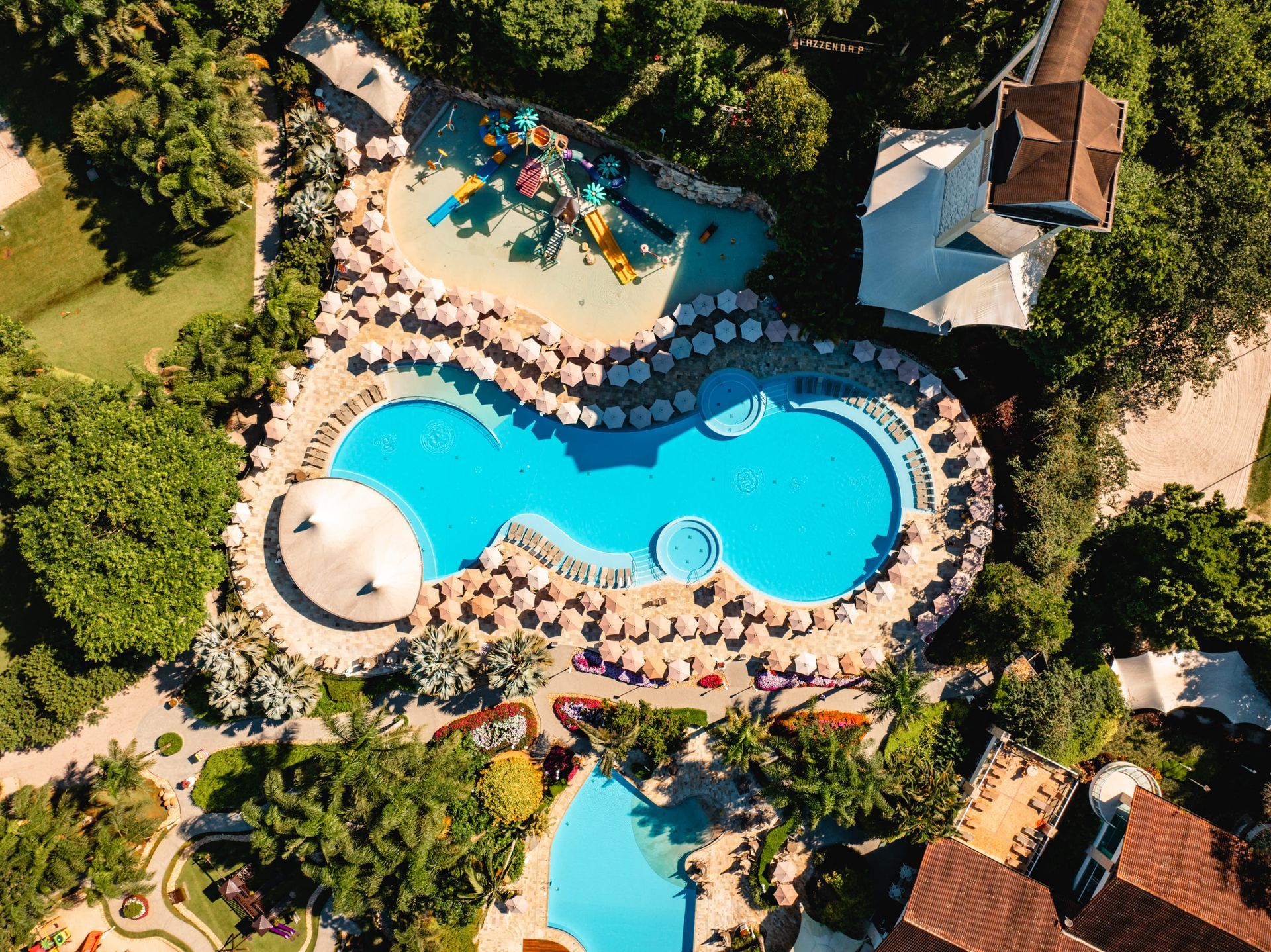 Vista aérea da piscina de um resort com água azul, espreguiçadeiras, palmeiras e edifícios circundantes.