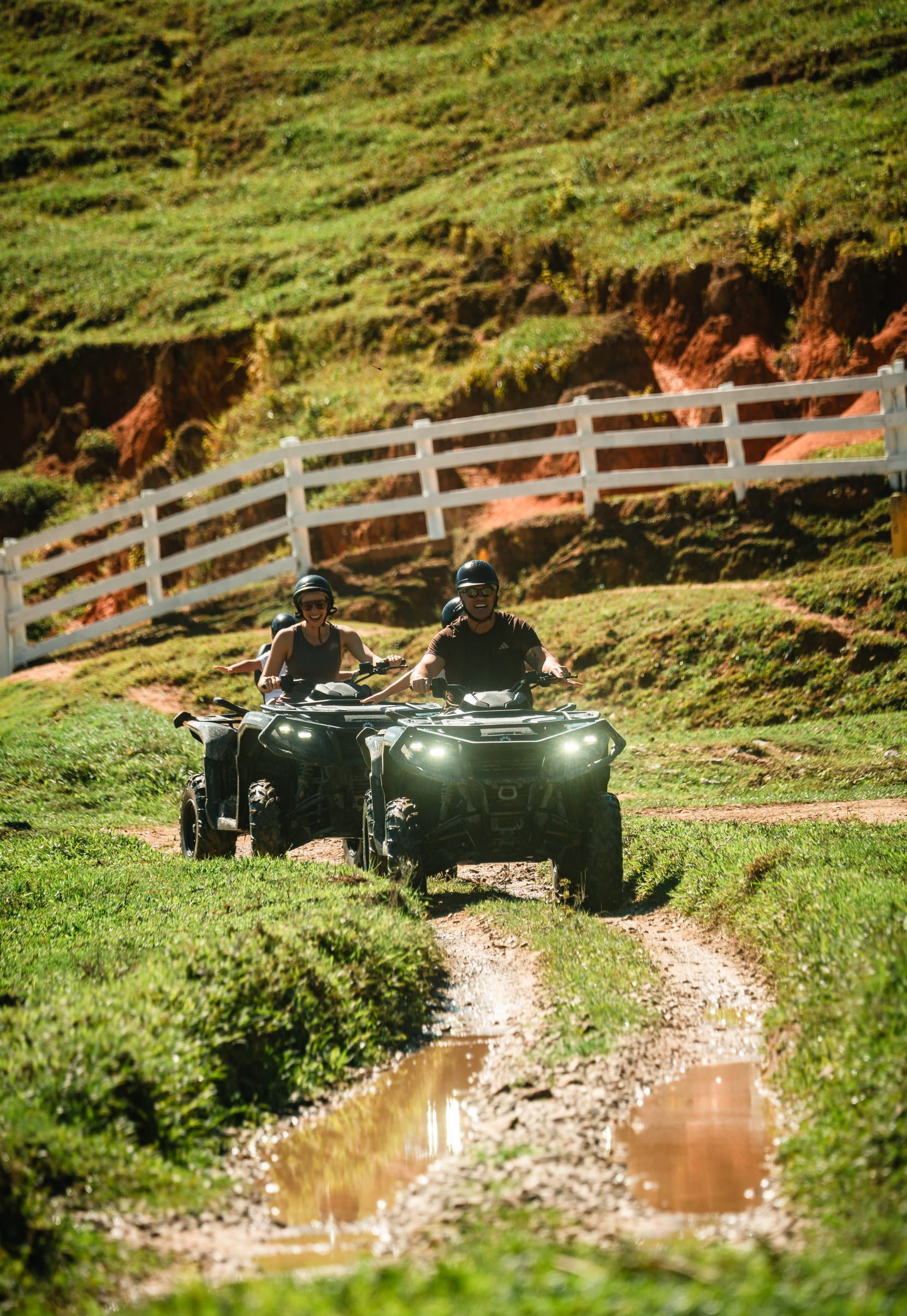 Duas pessoas conduzindo um quadriciclo em uma trilha lamacenta ao lado de uma cerca branca em um campo verde na encosta de uma colina.