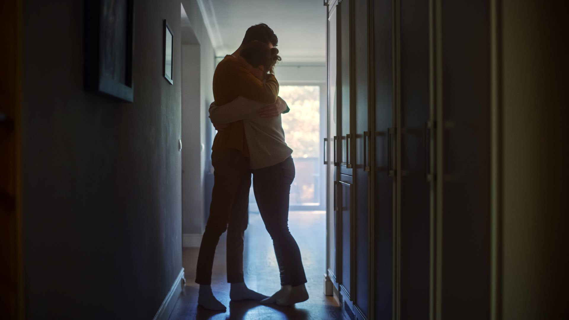 Two people embracing in a dim hallway; sunlight at the end, illuminating them.