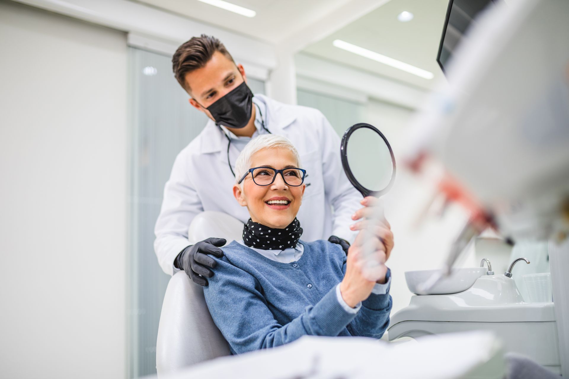 Dentist assisting a patient who is looking into a handheld mirror in the exam chair.