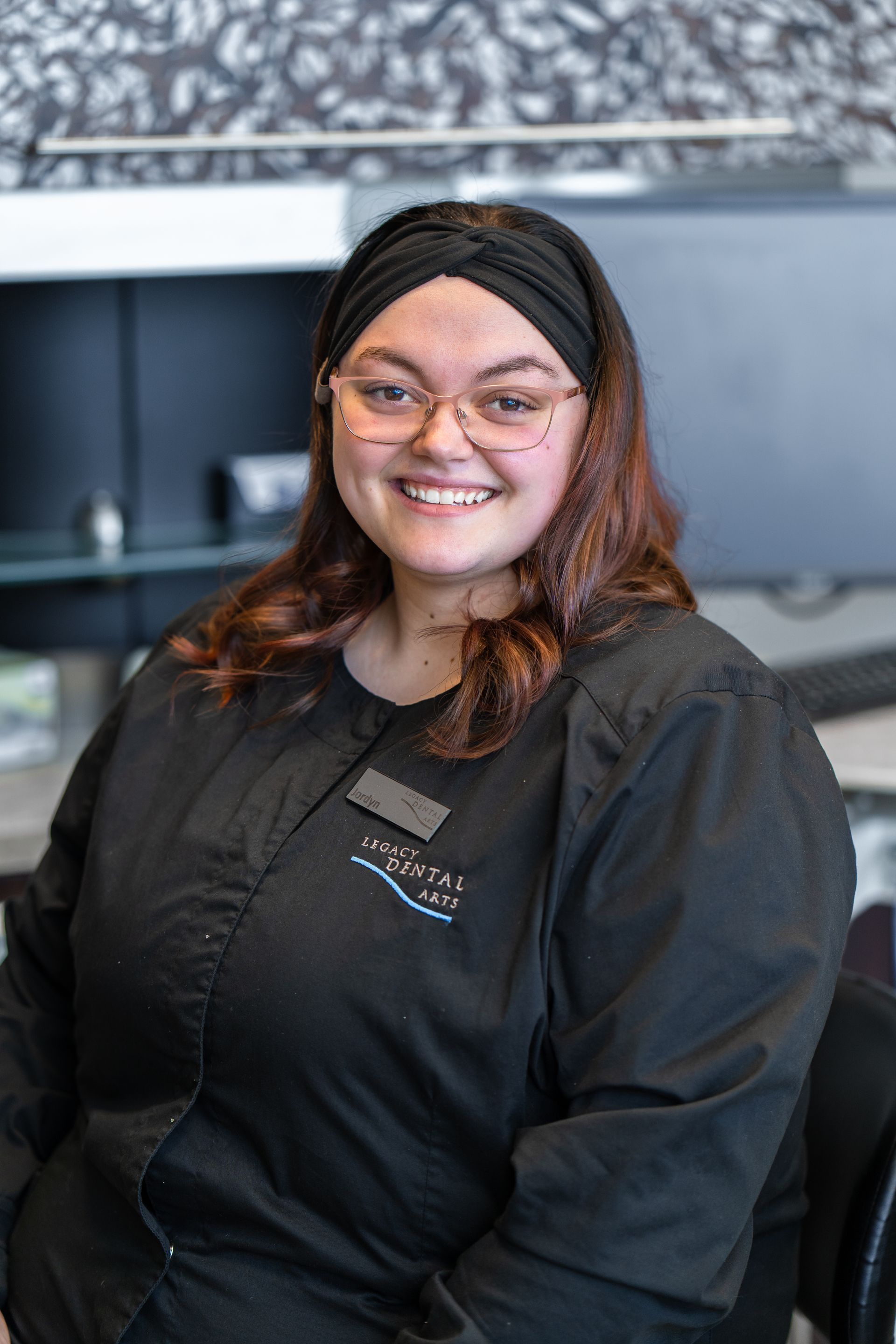 Woman wearing glasses and a black headband smiles, wearing a black shirt with a nametag, in an office setting.