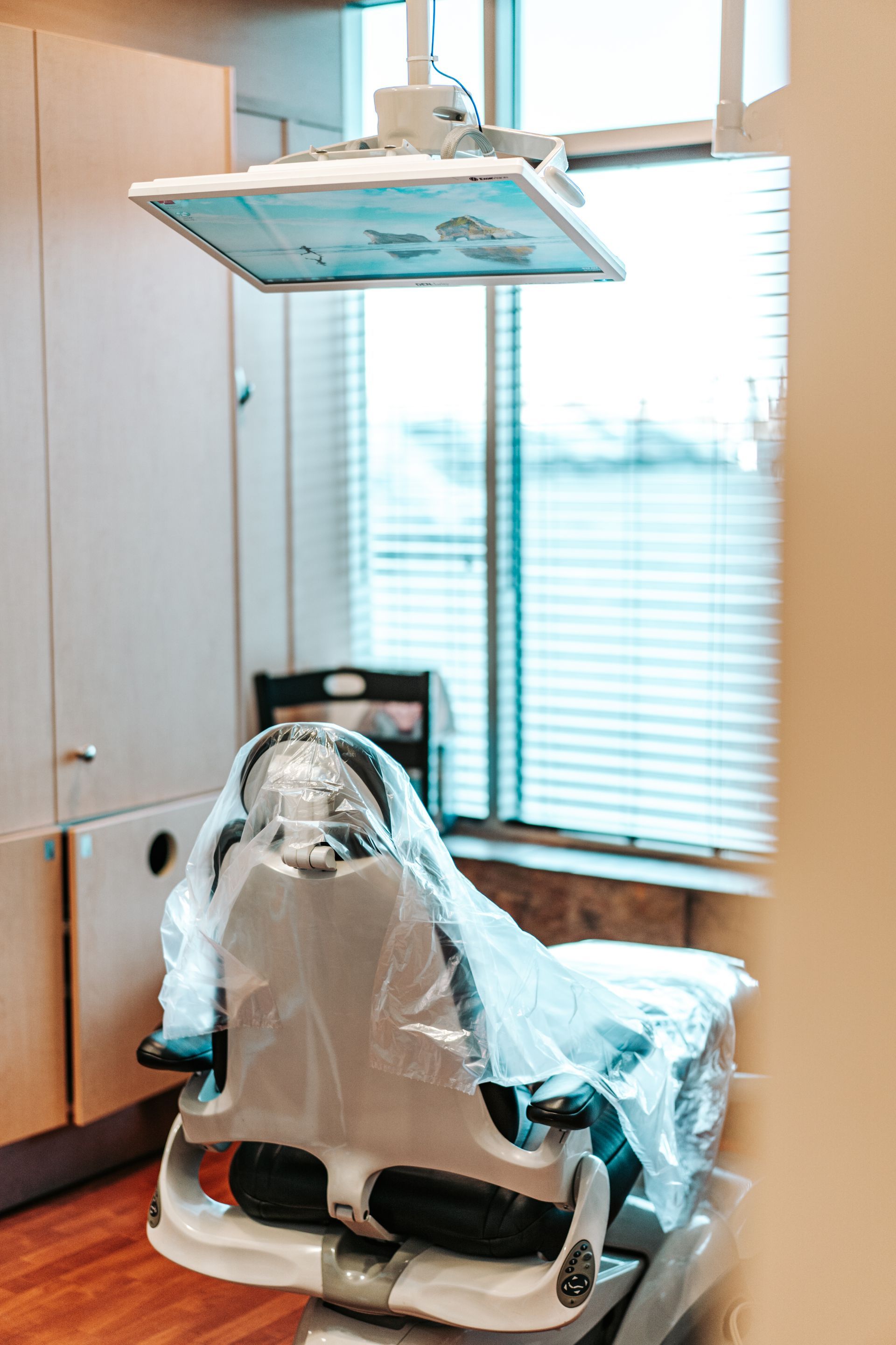 Dental chair covered in plastic, lit by overhead lamp, near a window with blinds.