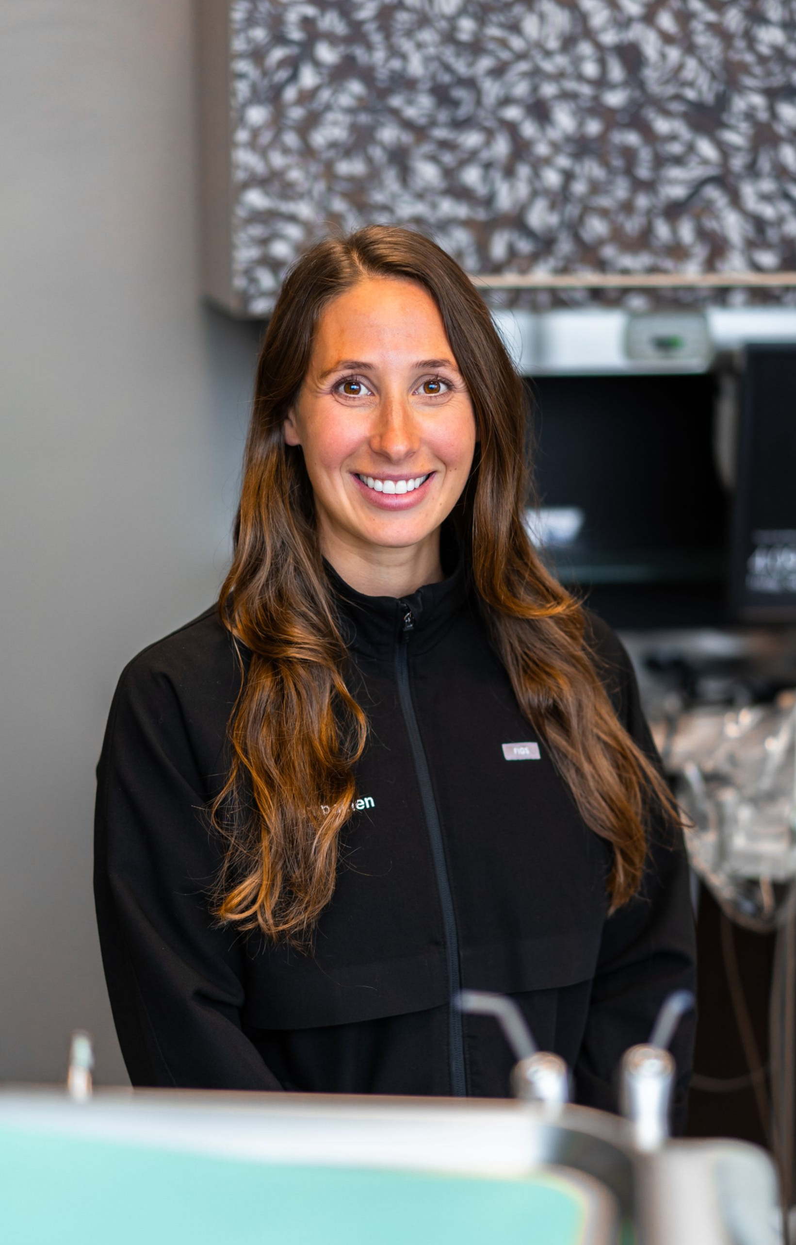 Woman with long brown hair smiles, wearing a black jacket, standing behind a counter with a patterned backdrop.