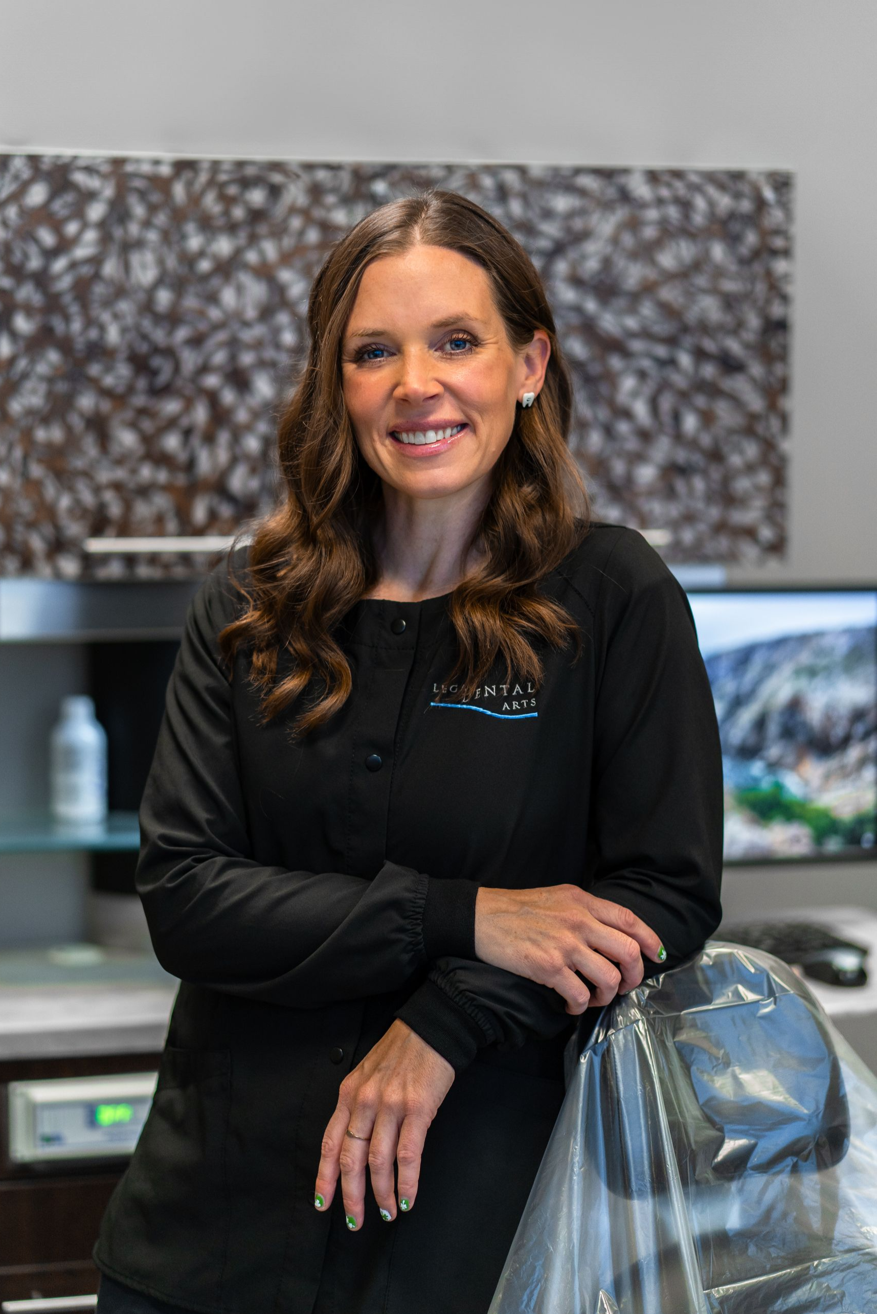 Woman in black scrubs, smiling, leans on dental chair in a modern dental office.