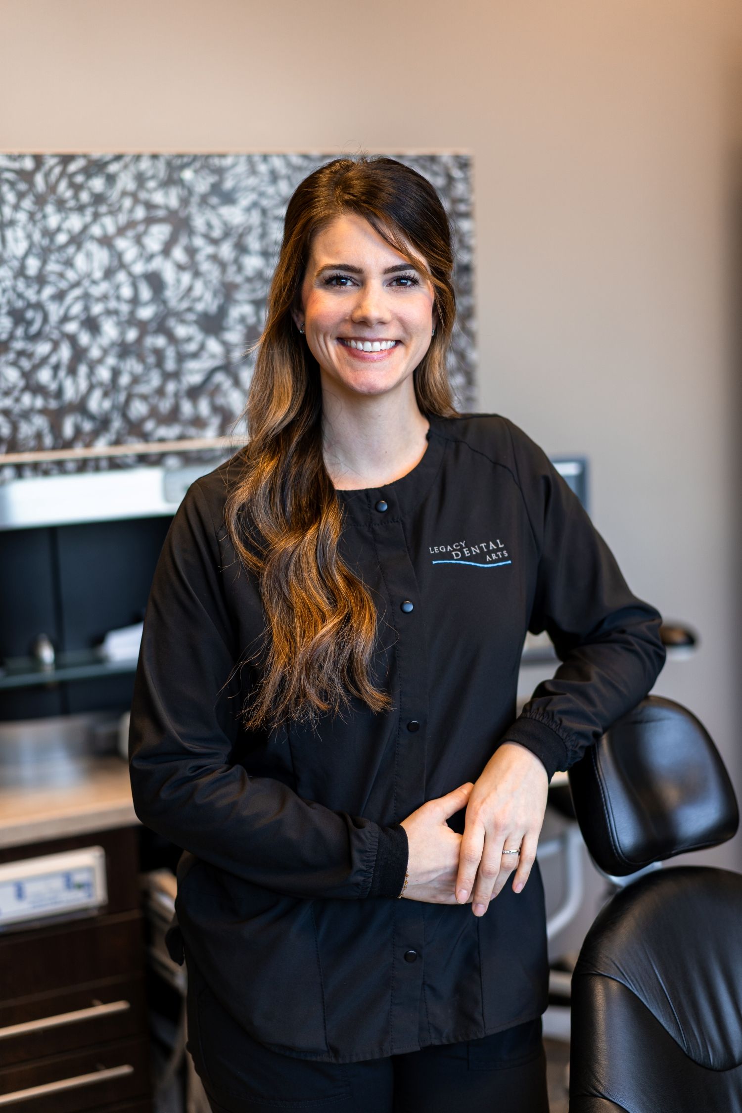 Smiling woman in black scrubs, leaning on dental chair in a dental office.
