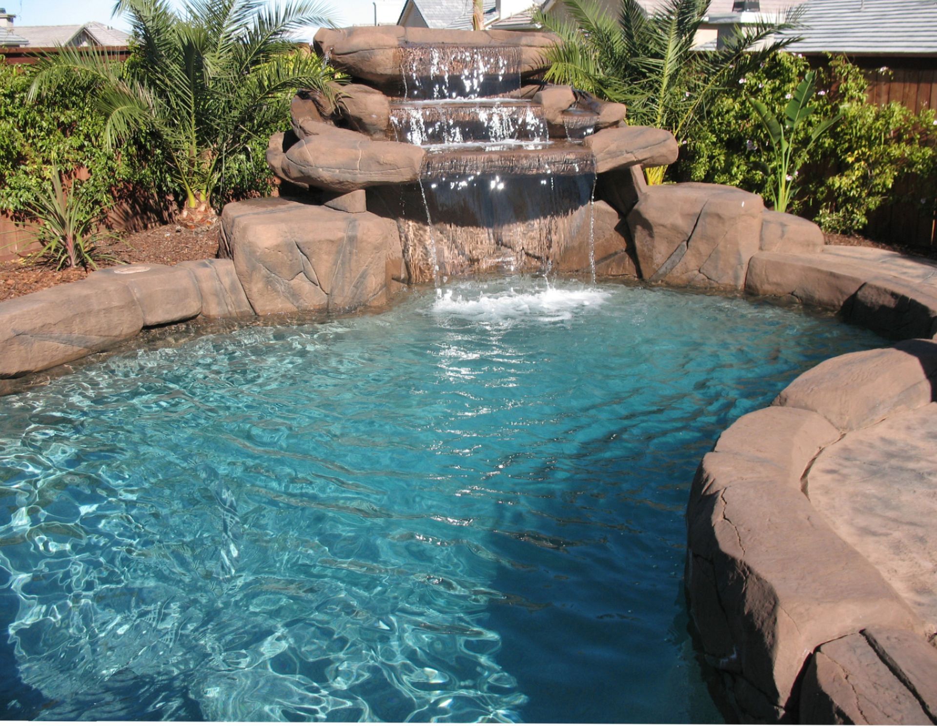 A three-tiered rock waterfall flowing into a blue-water swimming pool surrounded by lush green plants.