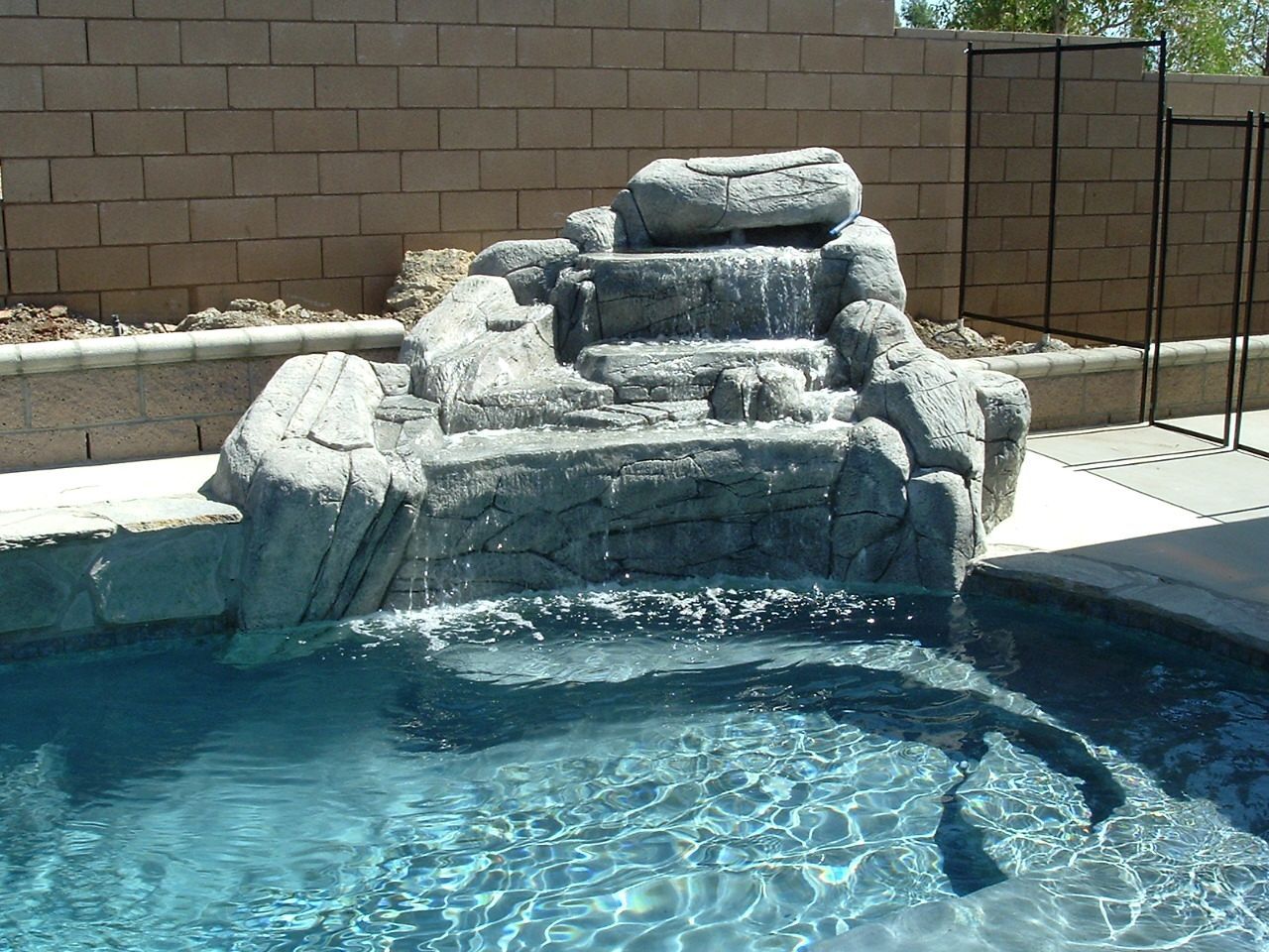 A faux-rock tiered waterfall feature flowing into a backyard swimming pool against a tan brick wall.