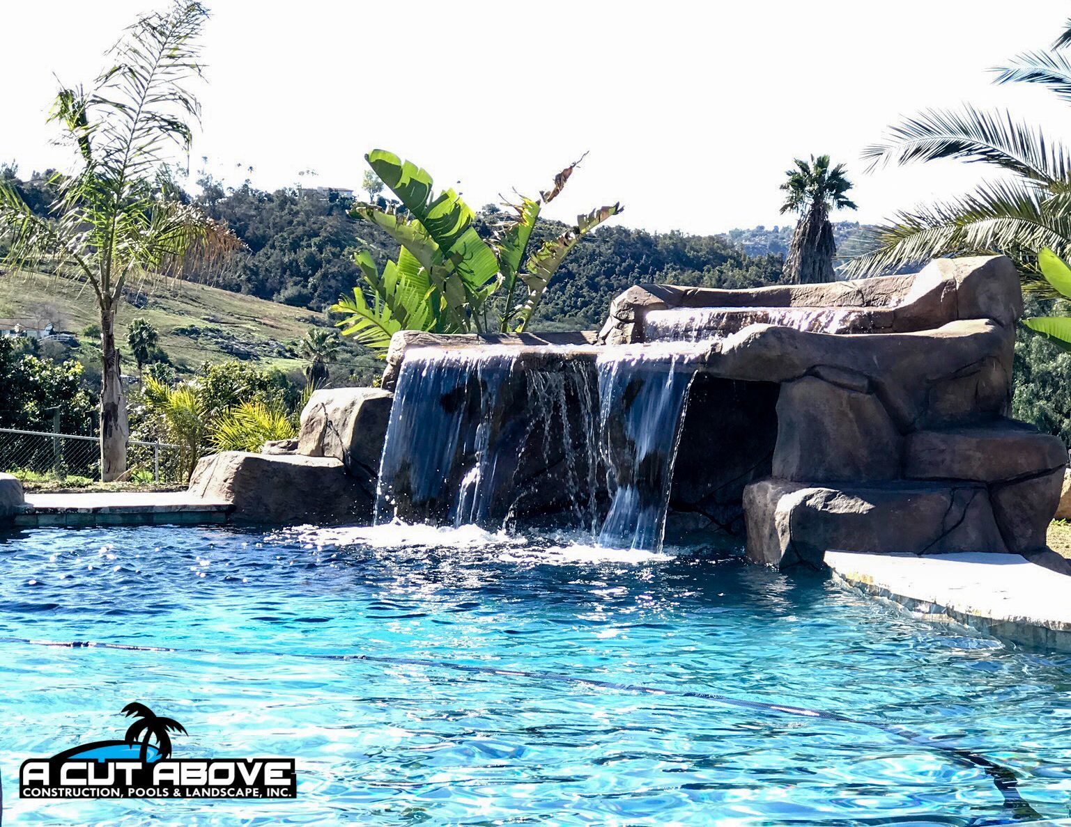 A multi-tiered artificial rock waterfall flowing into a blue swimming pool with a hilly, landscaped background.