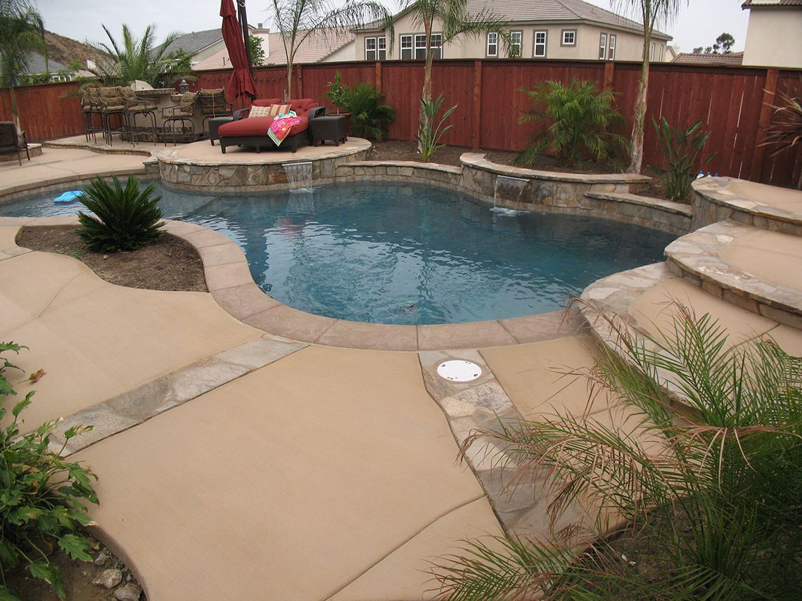 A backyard swimming pool with tan concrete decking, stone accents, a waterfall, and a privacy fence.