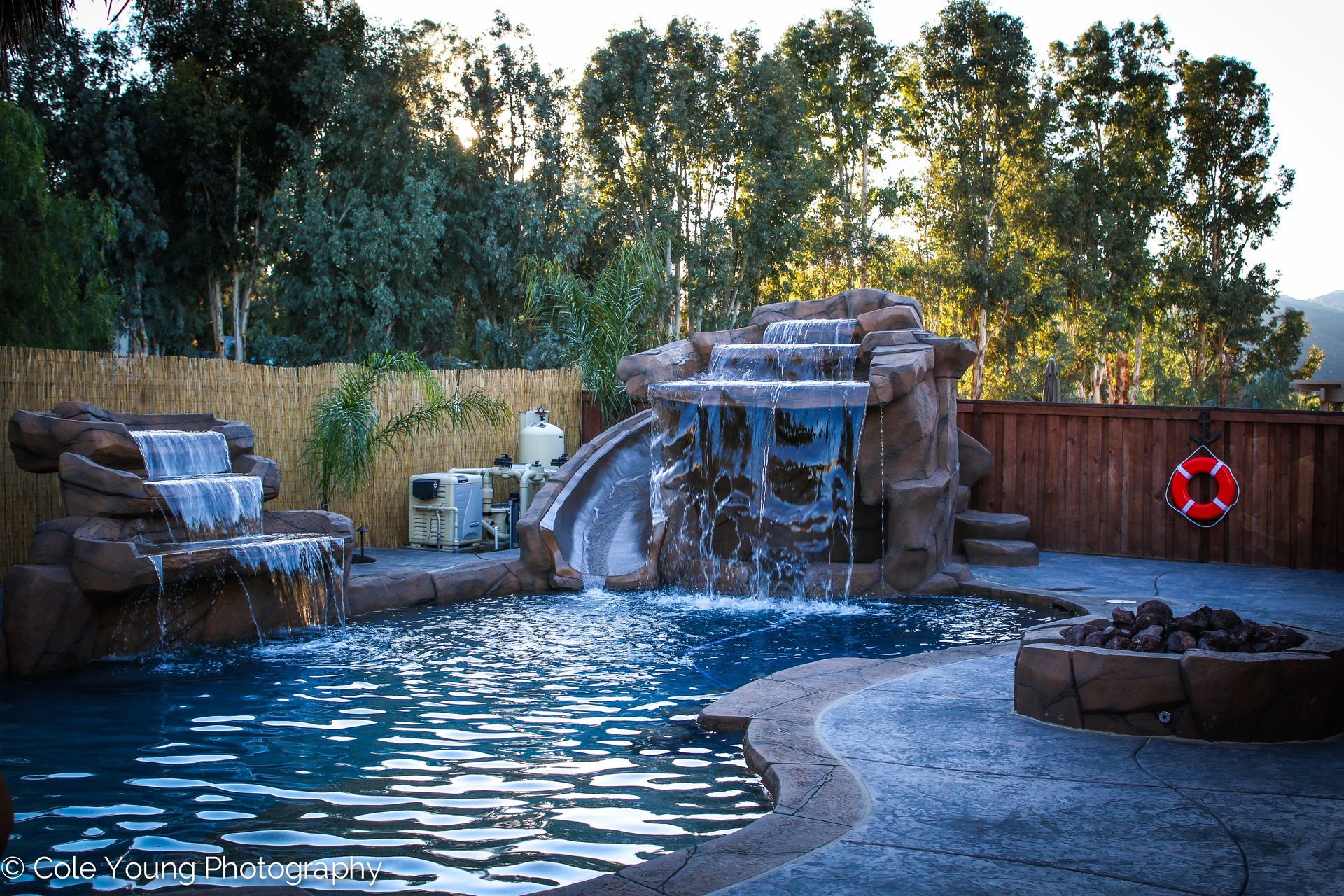 A swimming pool with two rock waterfalls, a slide, and a fire pit in a backyard with a wooden fence and trees.